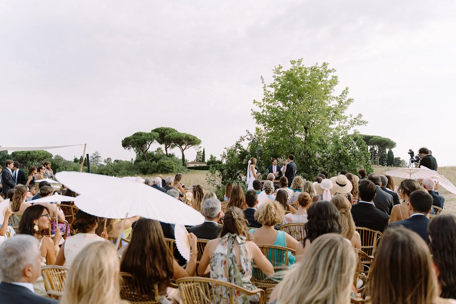 Outdoor wedding ceremony with guests seated under umbrellas, couples exchanging vows near a large tree, and the officiant officiating.