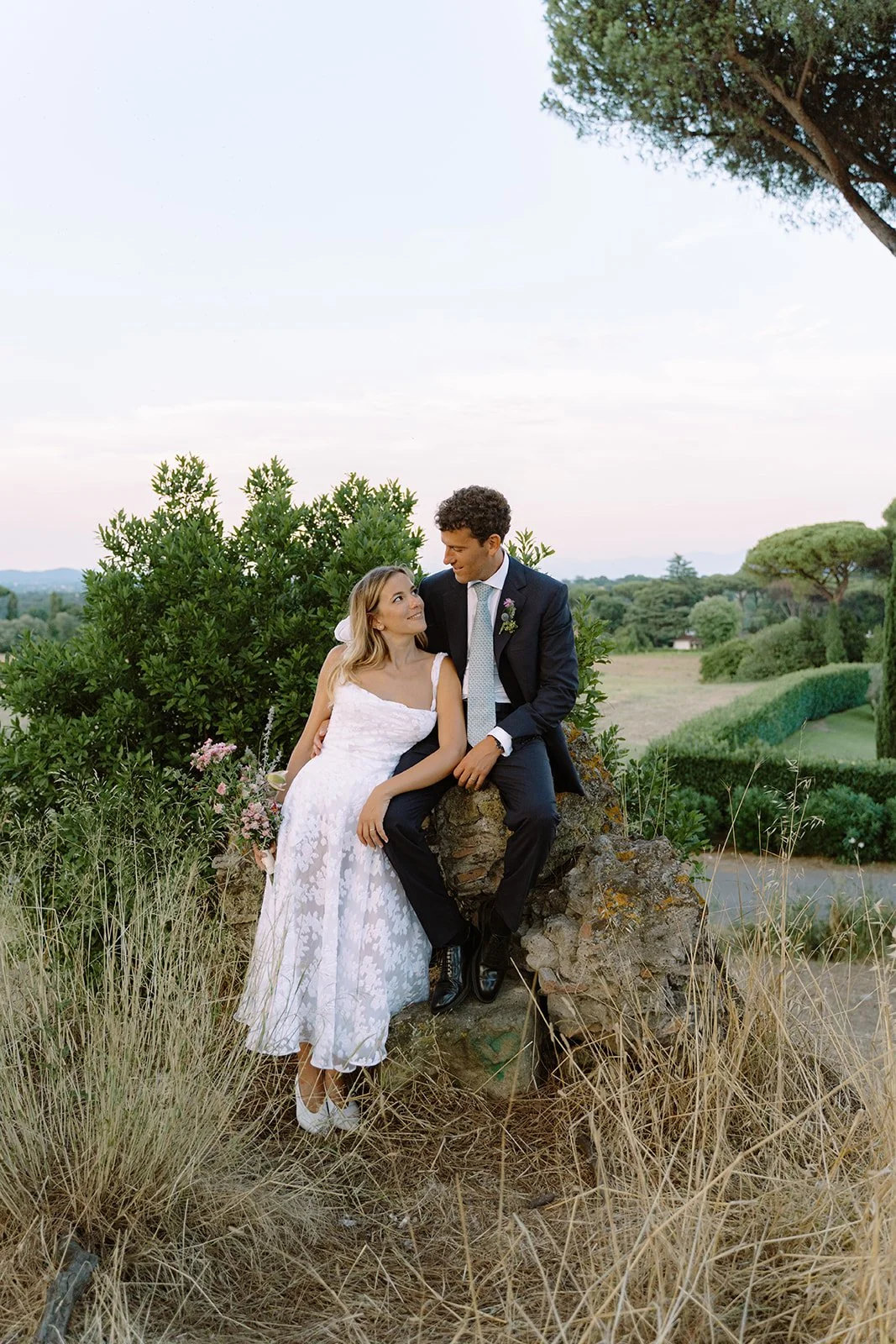 Bride and groom sitting on a rock outdoors, surrounded by greenery, looking at each other, during a wedding photoshoot at sunset.