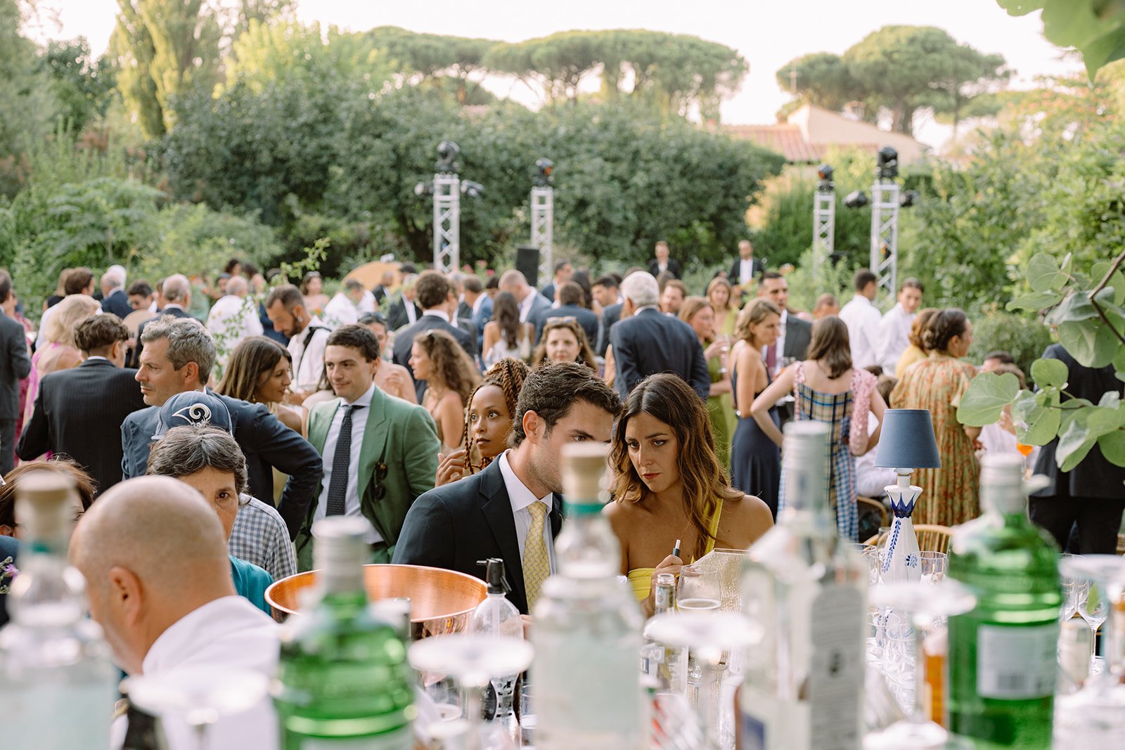 A crowd of people gathered outdoors at a social event, with some people talking and others looking on. There's a table in the foreground with bottles and a blue lamp. Tall trees and a building are in the background.