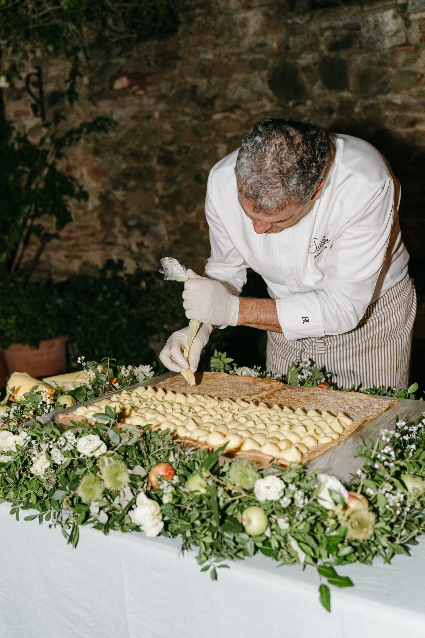 Chef decorating a large cake with cream, surrounded by white flowers and green foliage.
