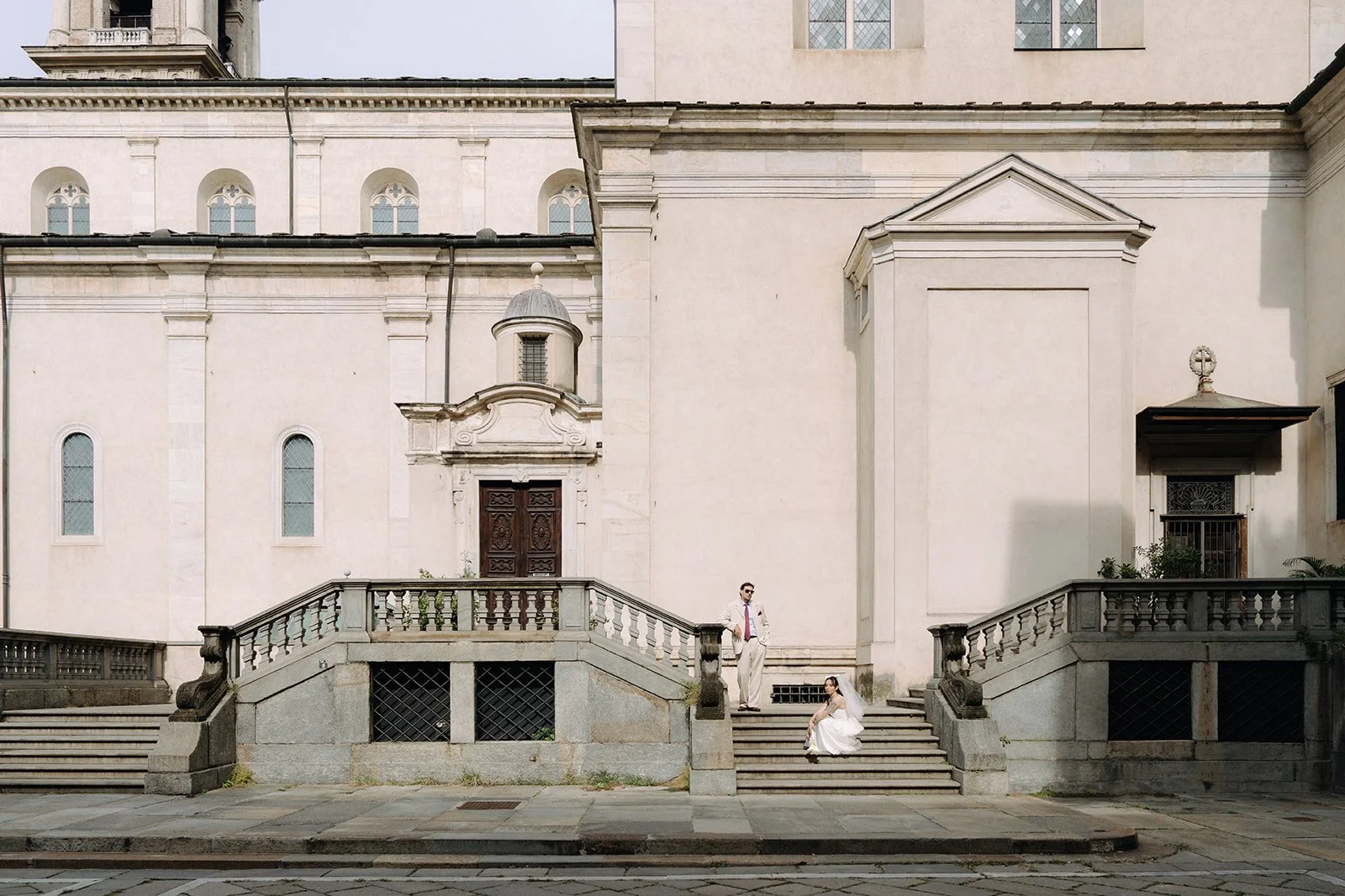 A bride in a white wedding dress sitting on steps and a groom in a white suit standing nearby outside a historic building with classical architecture.