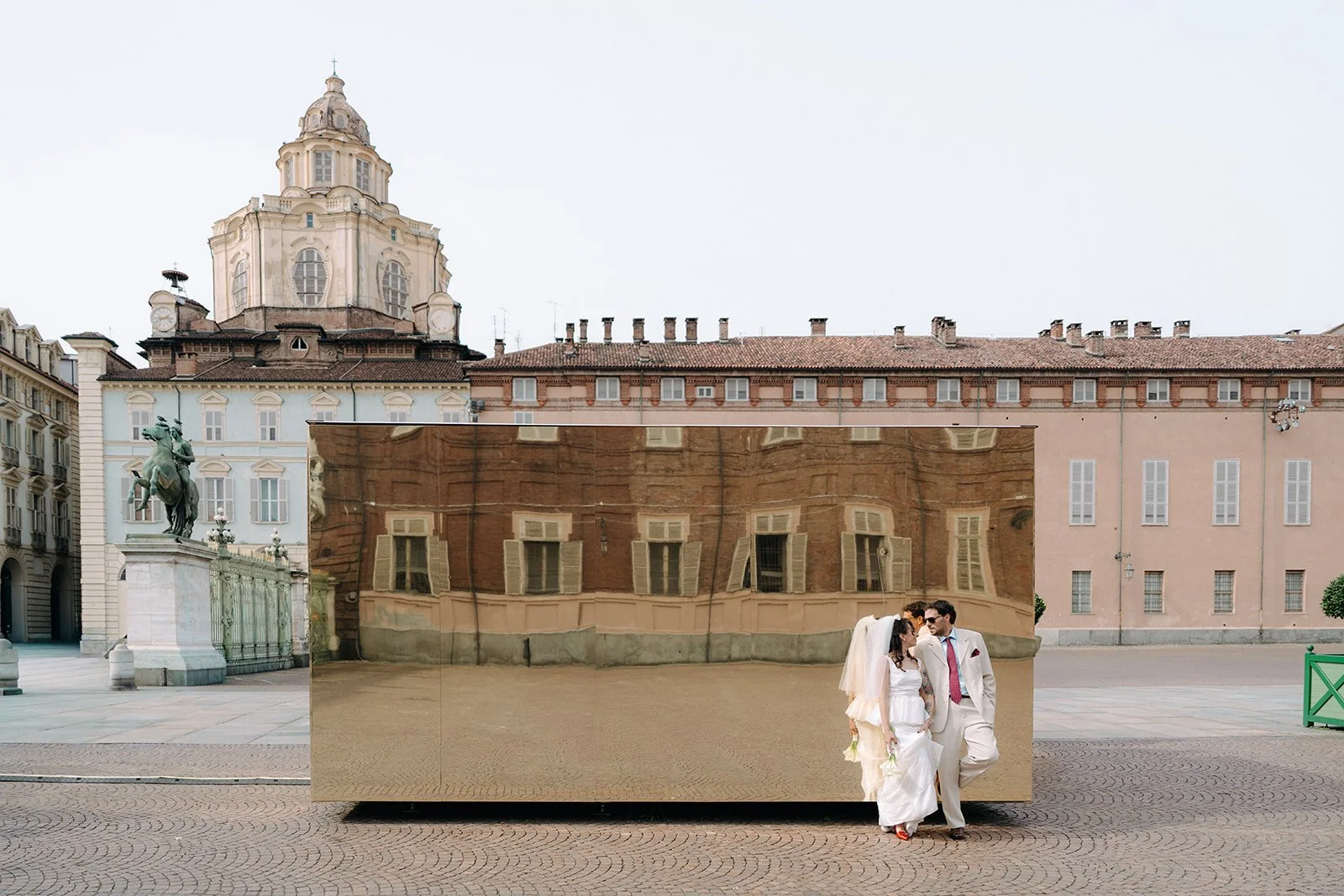 A couple dressed in wedding attire standing in front of a reflective, metallic sculpture in an urban square with historic buildings and a statue of a man on a horse in the background.
