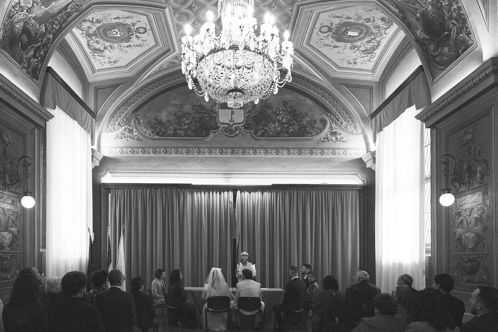 A wedding ceremony taking place in an ornate, historic room with high decorated ceilings, a large chandelier, tall curtains, and a group of people seated watching the bride and groom at the altar, with an officiant standing behind a table.