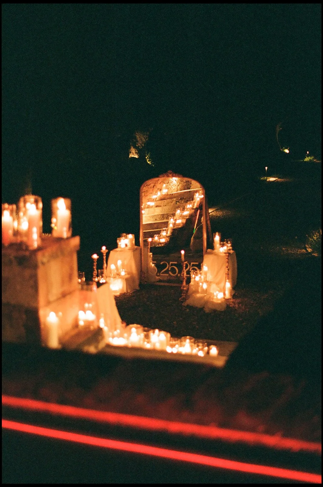 Memorial setup with numerous candles, a mirror, a photograph, and a sign with a date, suggesting a tribute or remembrance event at night.