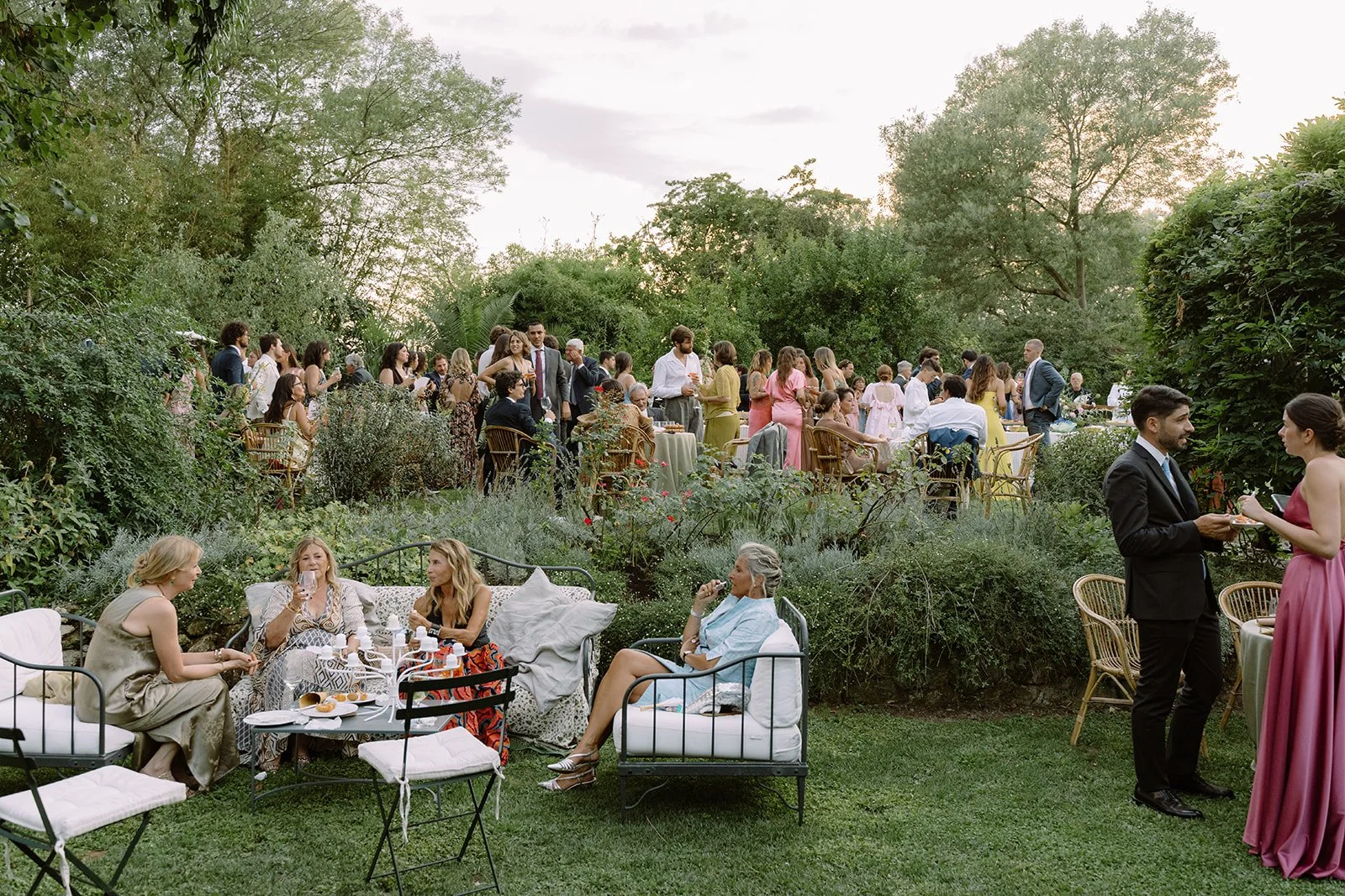 Guests enjoying an outdoor garden party with tables, chairs, and lush greenery during the evening. Some guests are seated and socializing, while others are standing and conversing.