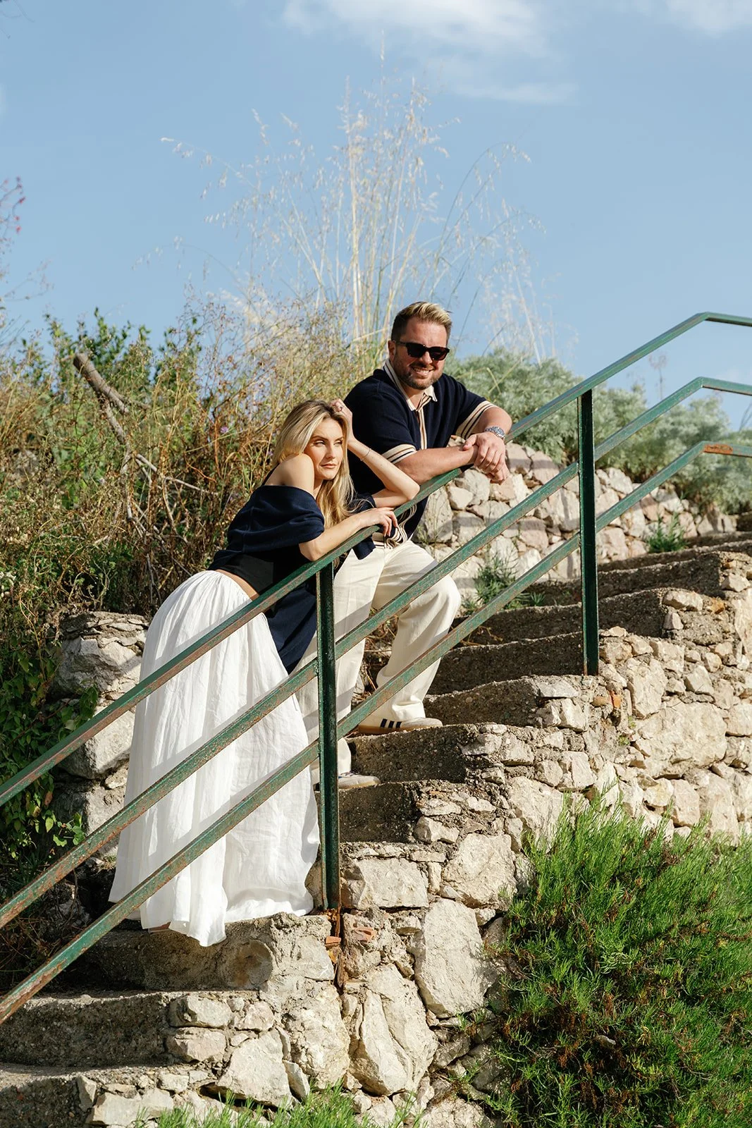 A man and woman pose on outdoor stone stairs with greenery and blue sky background. The woman is leaning on the railing, wearing a dark off-the-shoulder top and a long white skirt. The man, wearing sunglasses, a dark polo shirt, and light pants, is l