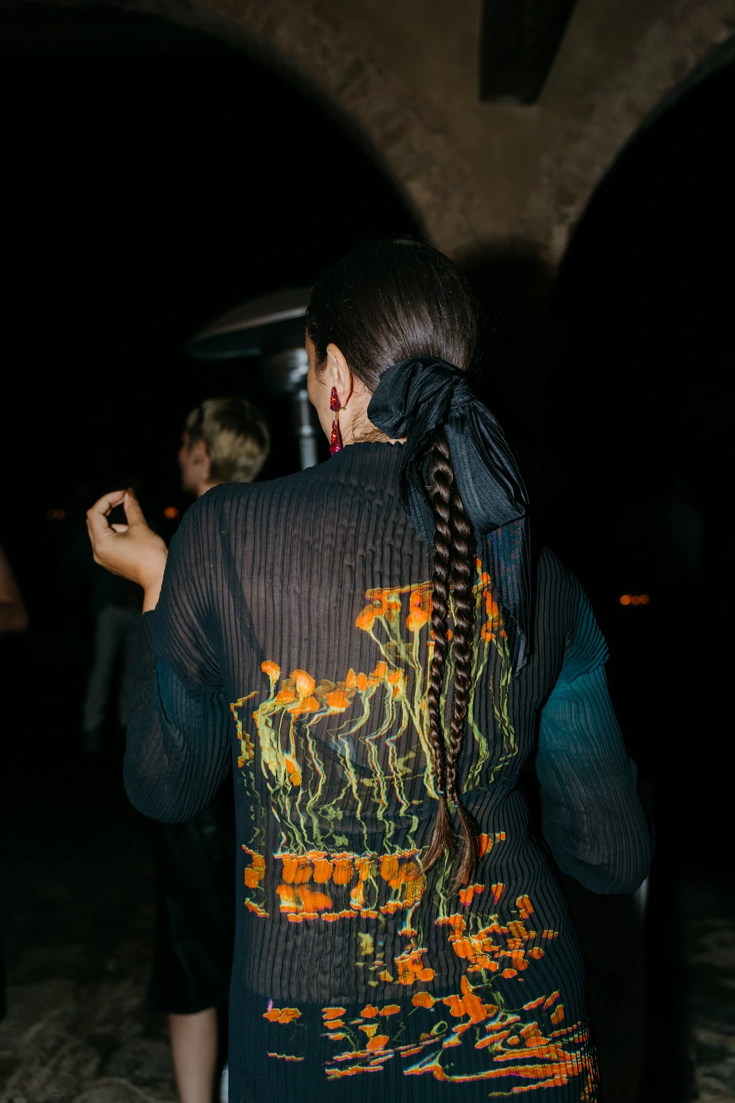 Back of a woman with long braided hair, black ribbon, red earrings, wearing a dark embroidered dress, at night.