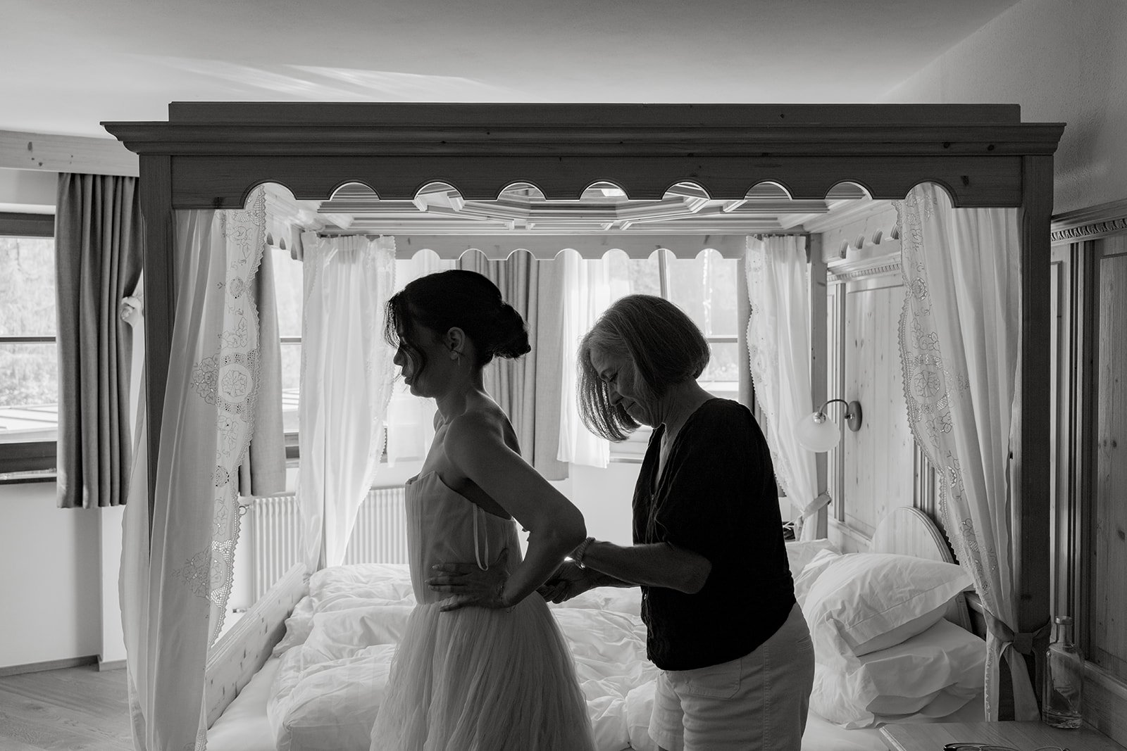 A woman in a dresses being helped by another woman in a bedroom with white curtains and a large bed.