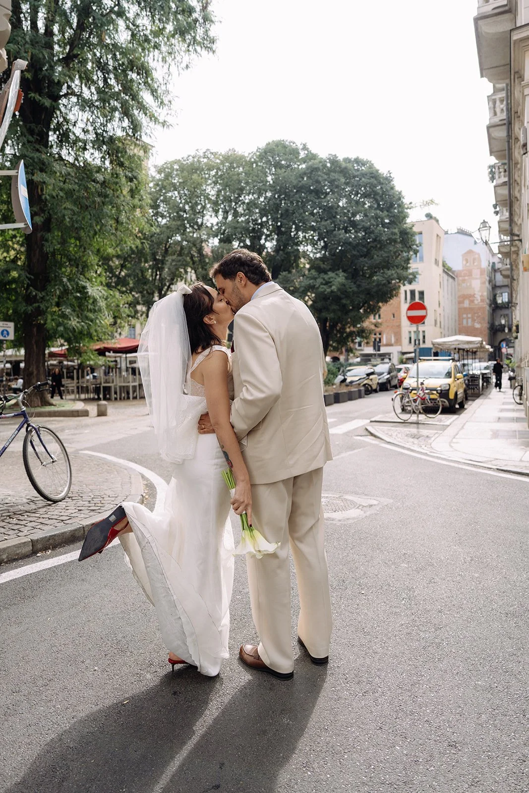 A bride and groom kissing on a city street; the bride lifts her right leg and holds a bouquet of white flowers; the groom wears a beige suit; trees and buildings are in the background.