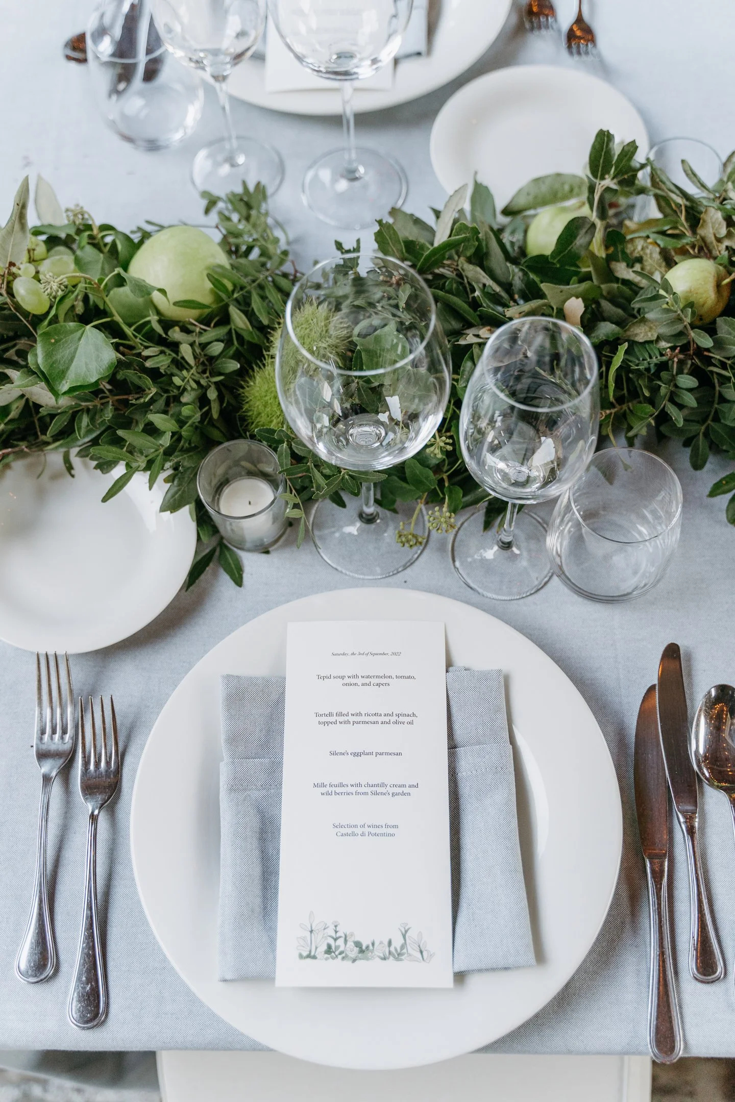 Elegant table setting with a white dinner plate, silverware, a folded napkin, a menu card with floral art, clear wine glasses, small candle holder, and a lush green floral centerpiece on a light grey tablecloth.