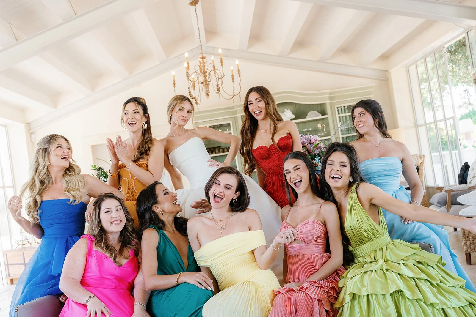 Group of women celebrating a wedding, with the bride in a white dress at center and bridesmaids in colorful dresses around her, in a bright, elegant room.