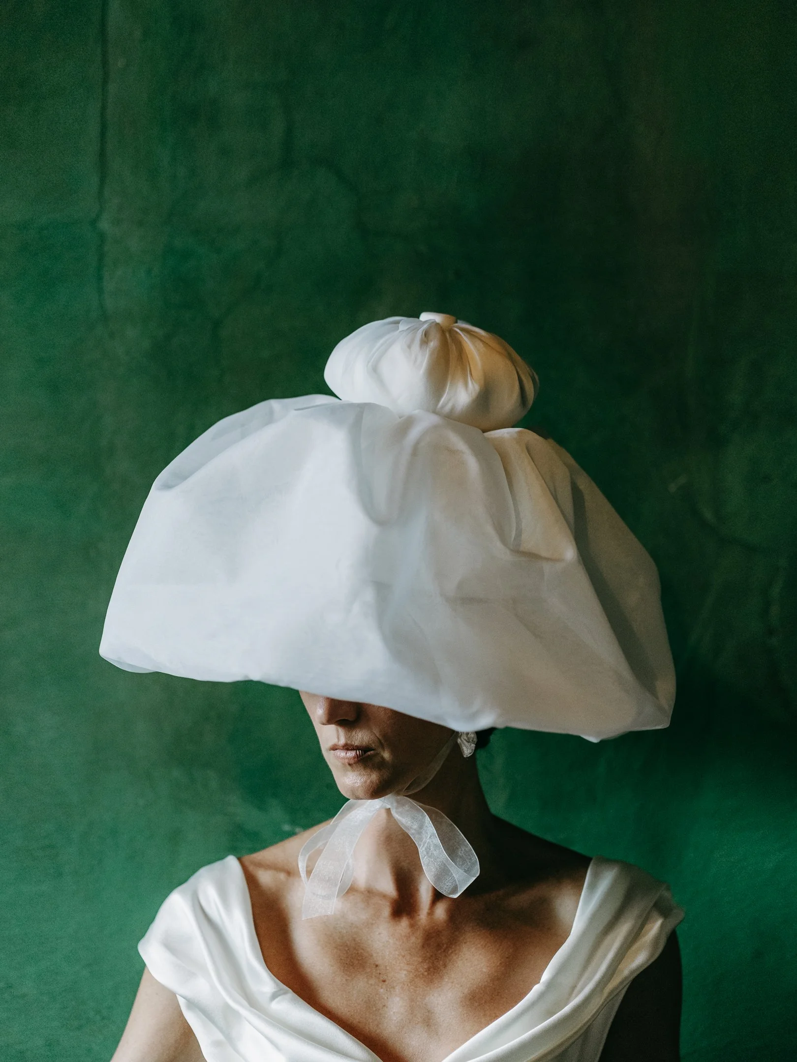 Woman wearing an oversized white hat with a small white bag on top, a white satin dress, and earrings against a green textured background.