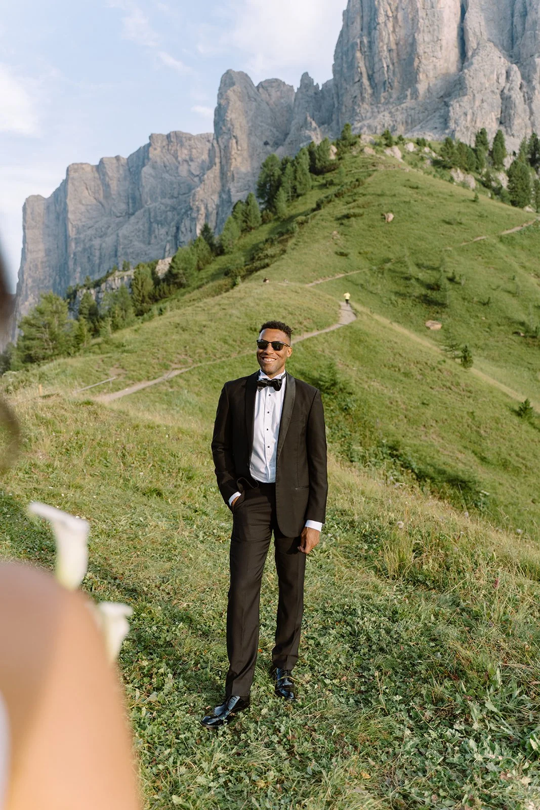 A man in a black tuxedo with a bow tie, sunglasses, and open collar shirt standing outdoors in front of green hills and rocky mountains, smiling.