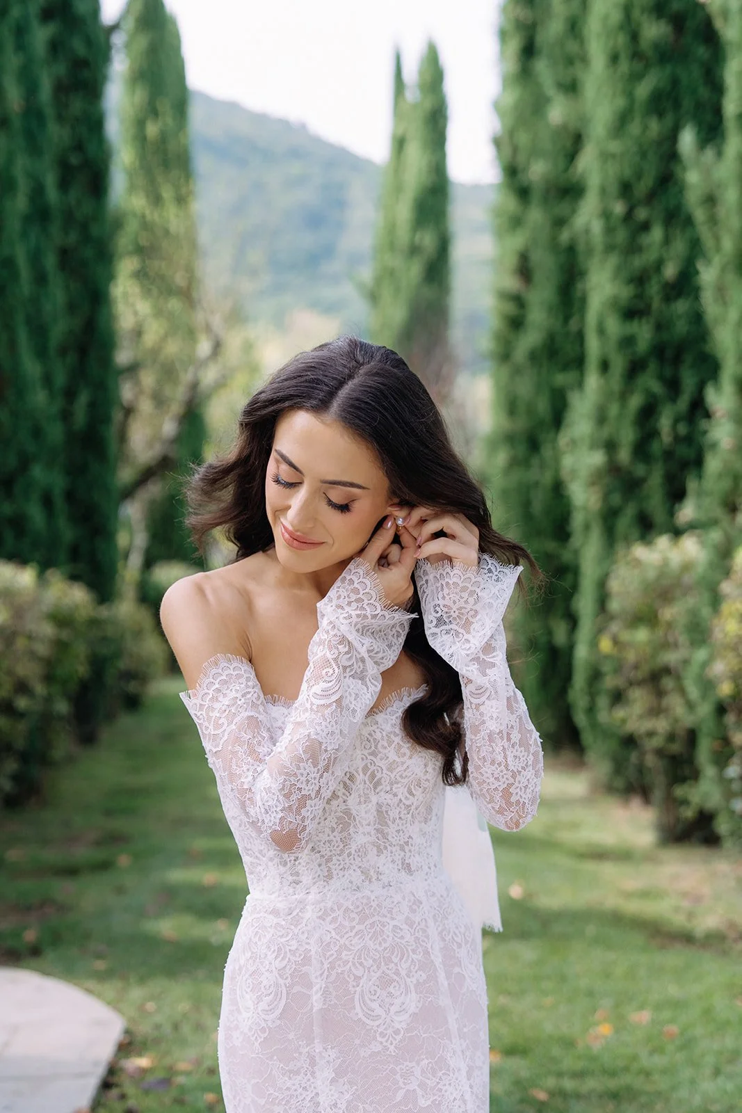 A woman in a white lace off-the-shoulder dress is adjusting her earring outdoors, surrounded by tall green cypress trees and lush greenery.