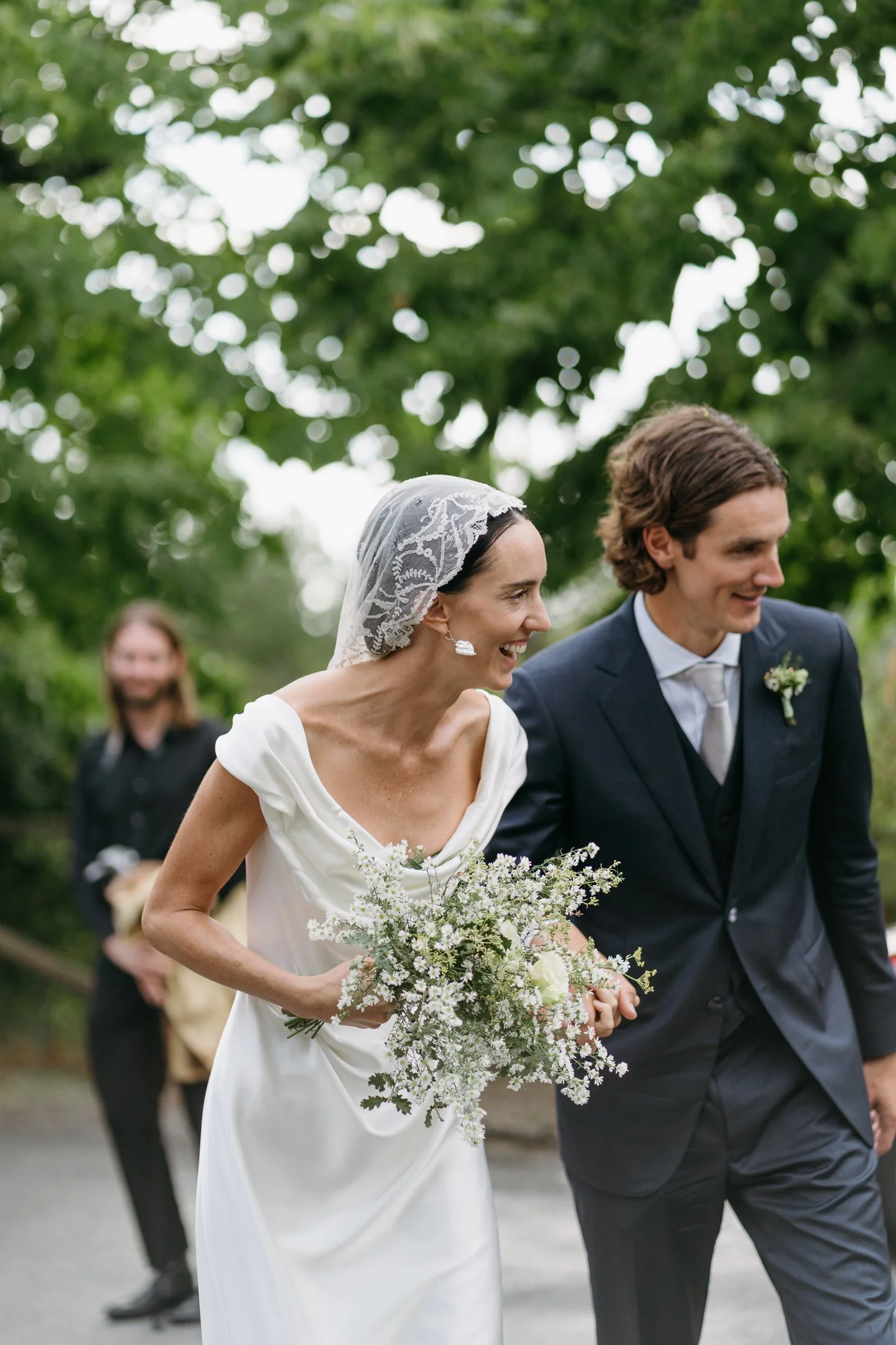 A bride and groom smiling and holding hands during their outdoor wedding, with a person in the background holding a dog.