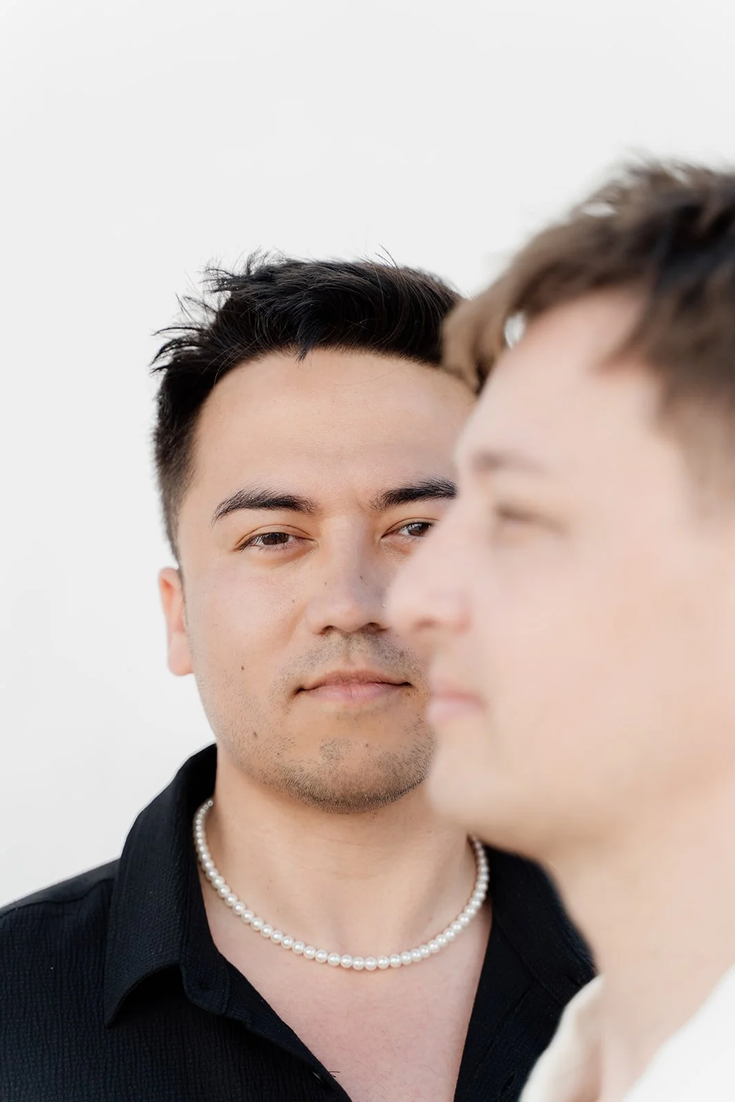 Close-up of two people, one Jamaican man with dark hair wearing a pearl necklace, and another person blurred in the foreground, both with neutral expressions against a plain white background.