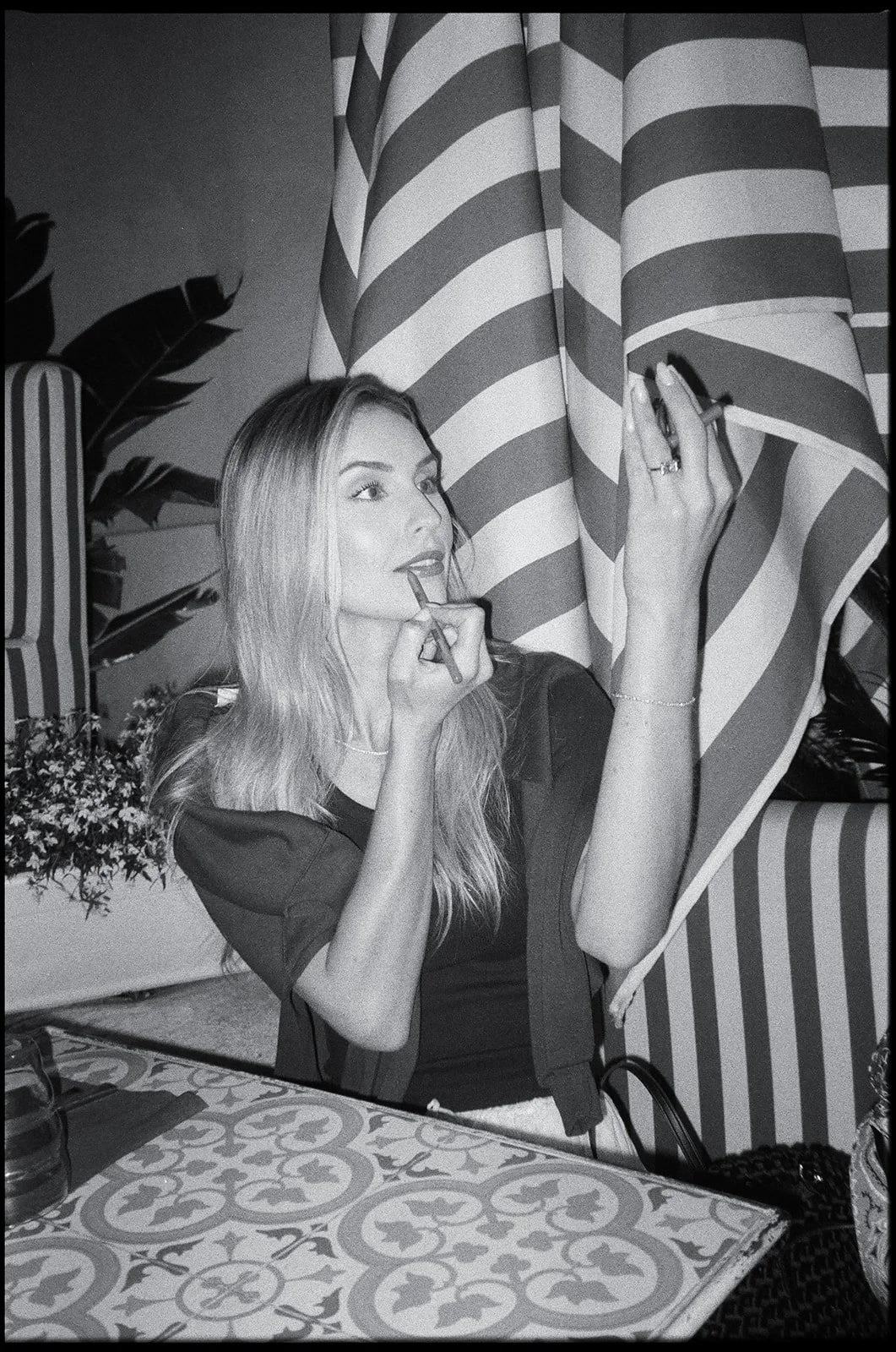 A woman sitting at a table, applying lipstick in front of a striped curtain, with decorative plants in the background.