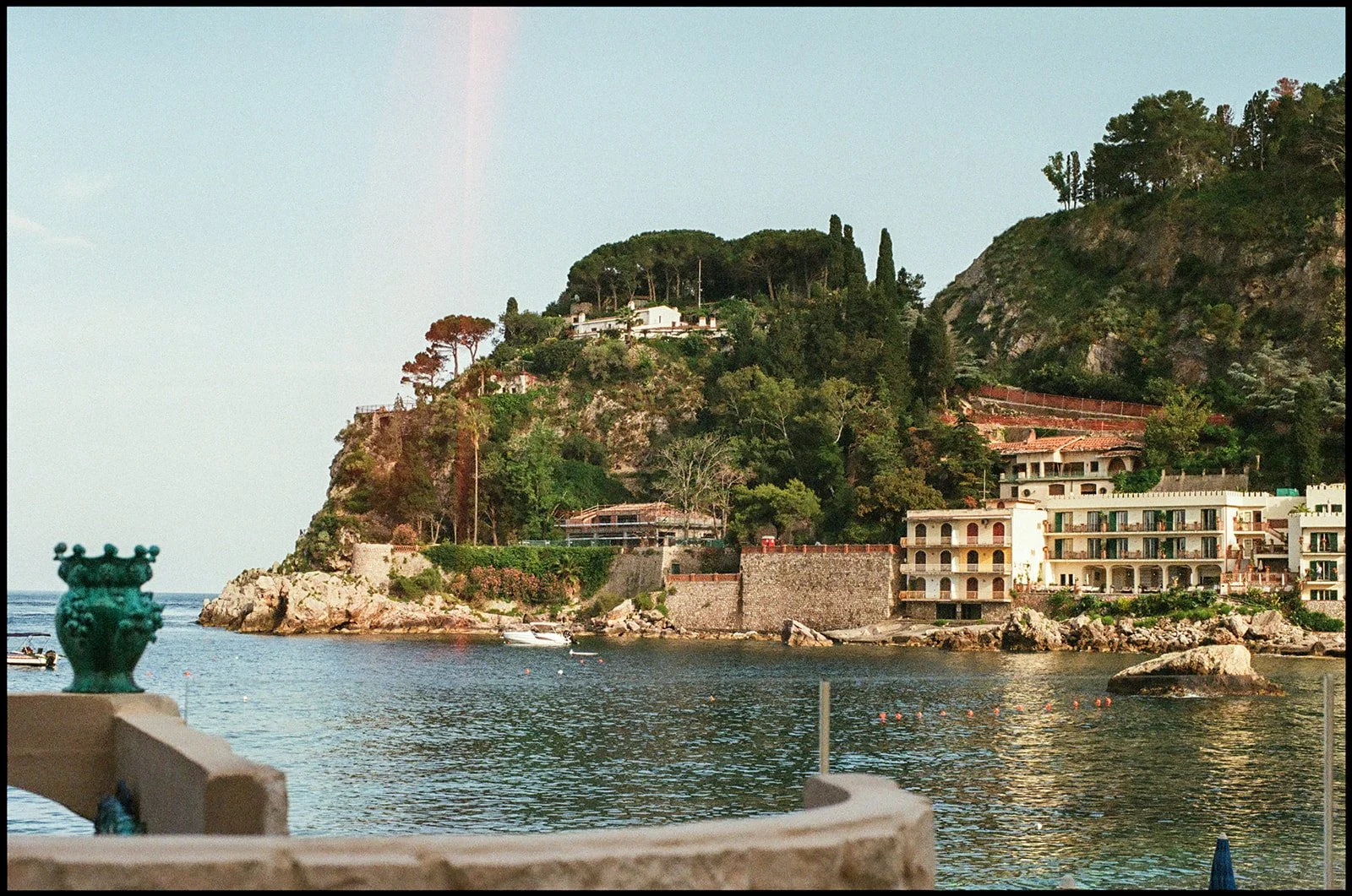 A scenic view of a coastal hillside with buildings, trees, and rocky formations, seen from a waterfront with water, boats, and an ornate green statue in the foreground.