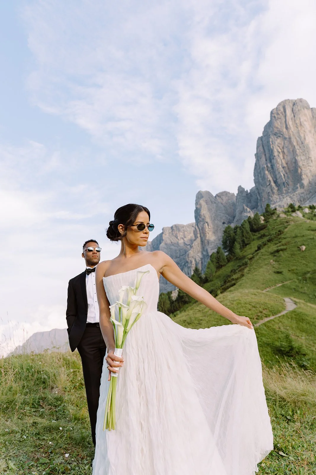 A bride in a white dress holding calla lilies in an outdoor mountainous landscape, with a groom in a black tuxedo standing behind her.