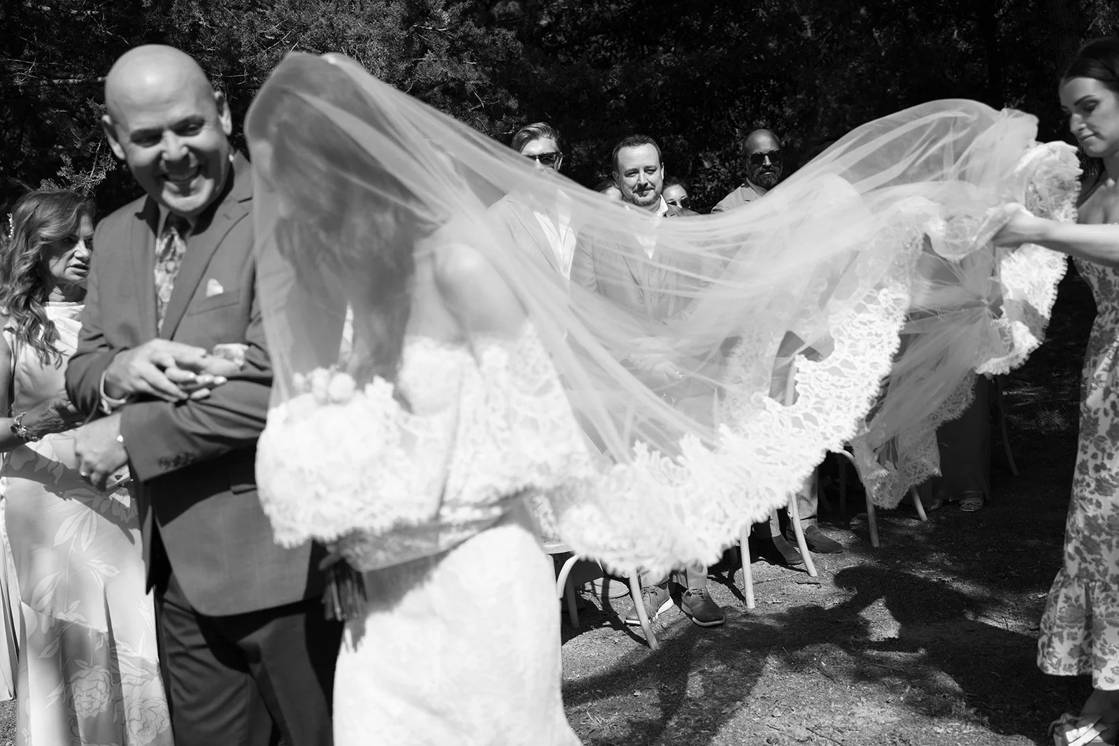 Black and white photo of a wedding scene with a bride in a lace gown and veil, surrounded by guests. The bride is lifting her veil, revealing her face in front of seated guests and other wedding party members, all outdoors in a sunny setting.