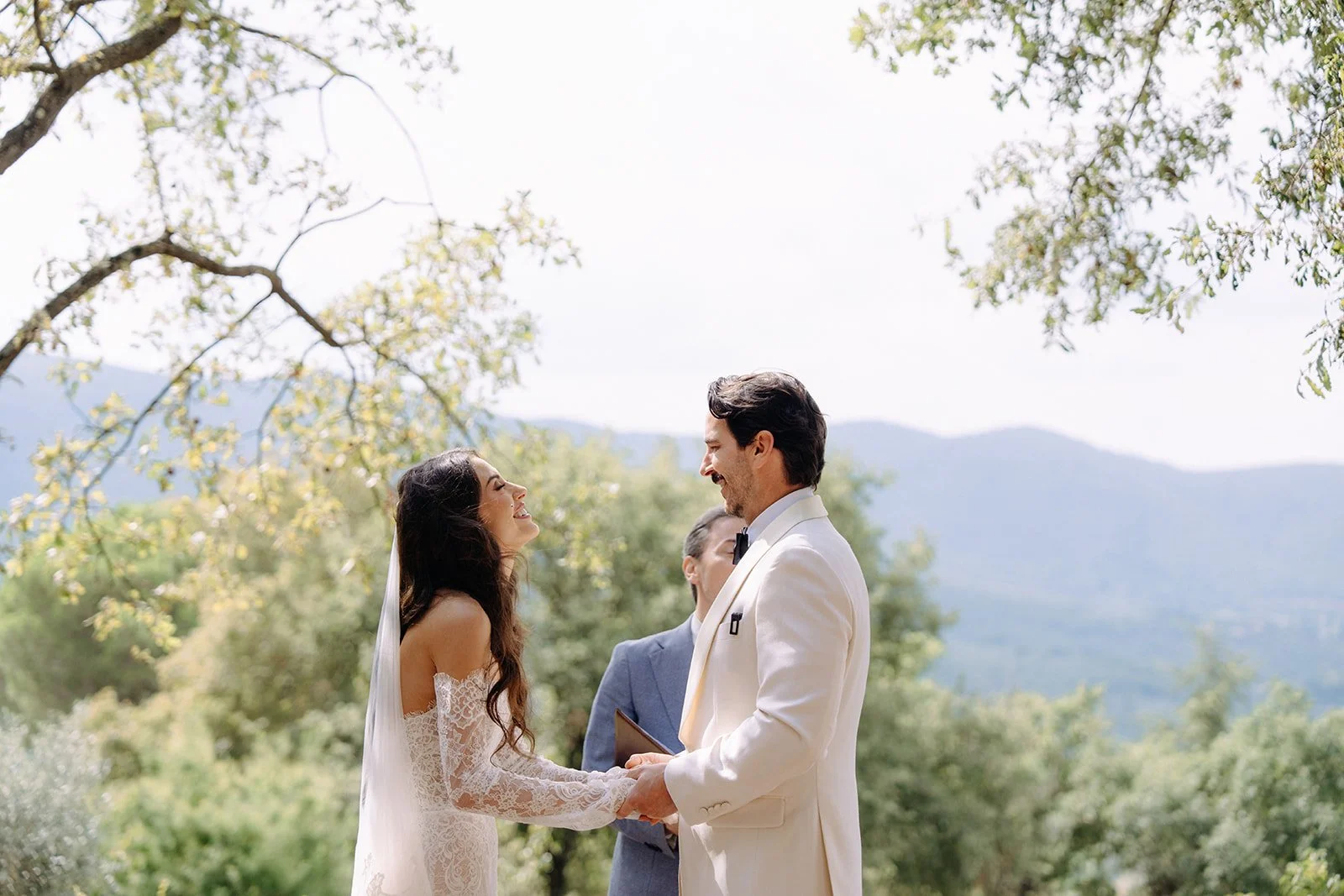 A couple in wedding attire exchanging vows outdoors, holding hands, with officiant in background, surrounded by trees and mountains.
