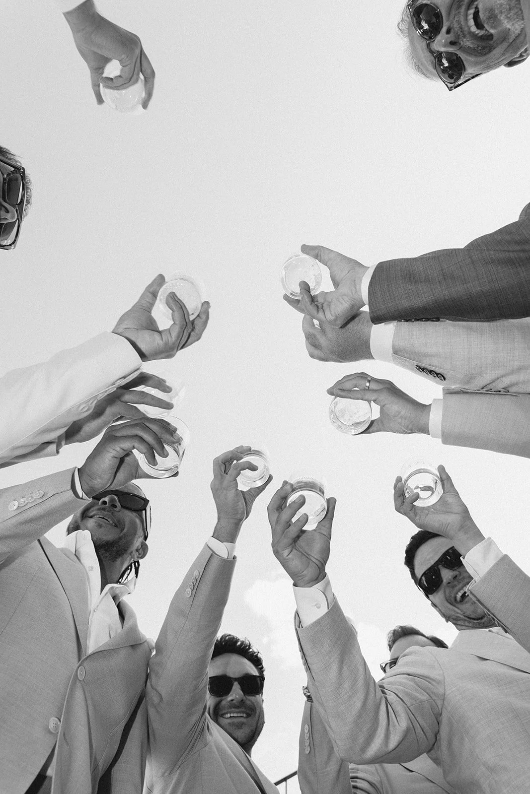 Group of men in suits wearing sunglasses, raising glasses in a toast, photographed from below against sky