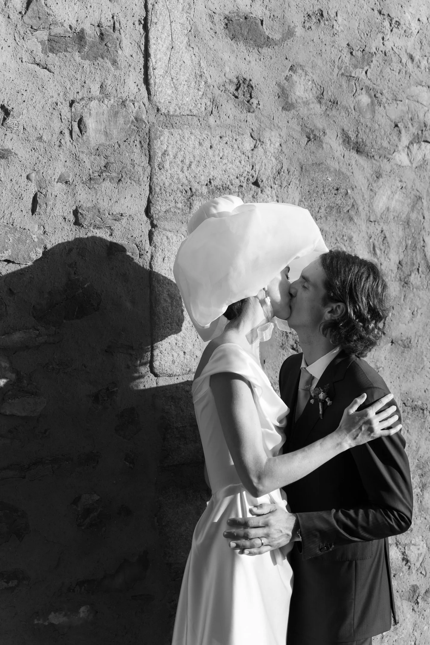 A black-and-white photo of a couple kissing, with a stone wall in the background. The woman is wearing a hat and a dress, and the man is in a suit. Their shadows are cast on the wall.