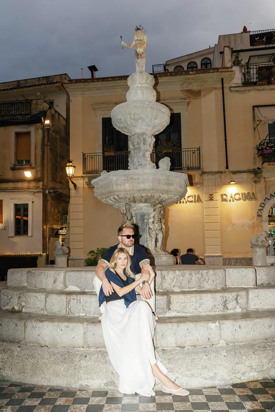 A man and woman sitting together in front of a large ornate stone fountain in a European city square during evening, with buildings and streetlights in the background.