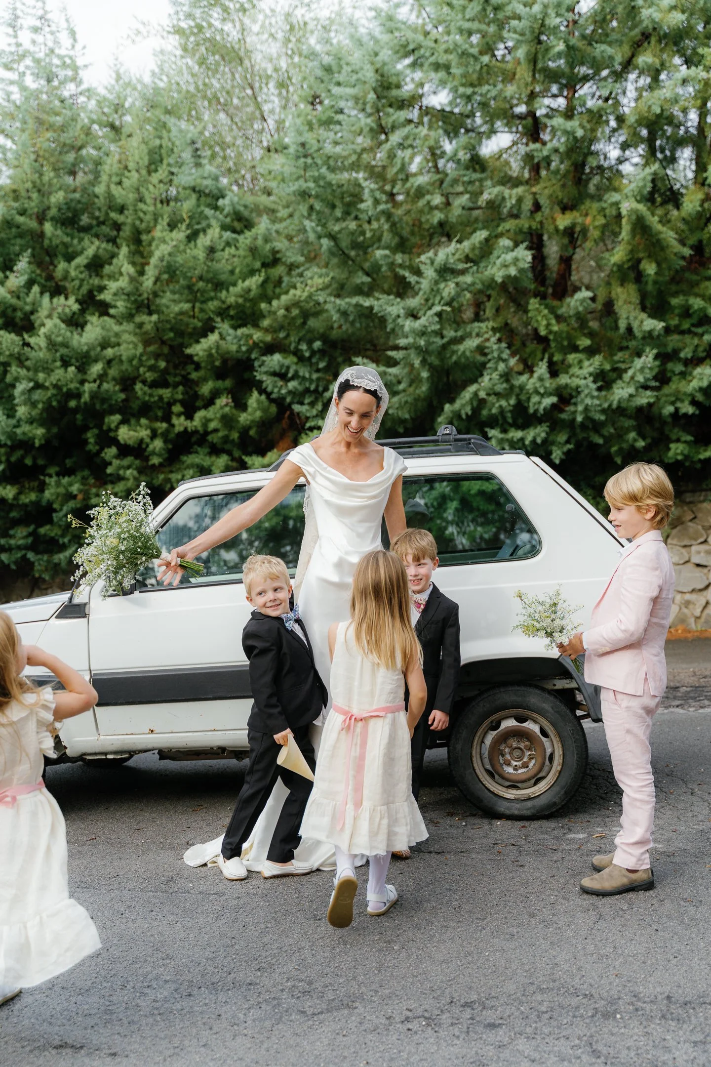 A bride in a white dress and veil stands by a white car, holding a bouquet of flowers, surrounded by children dressed in formal attire, with greenery in the background.