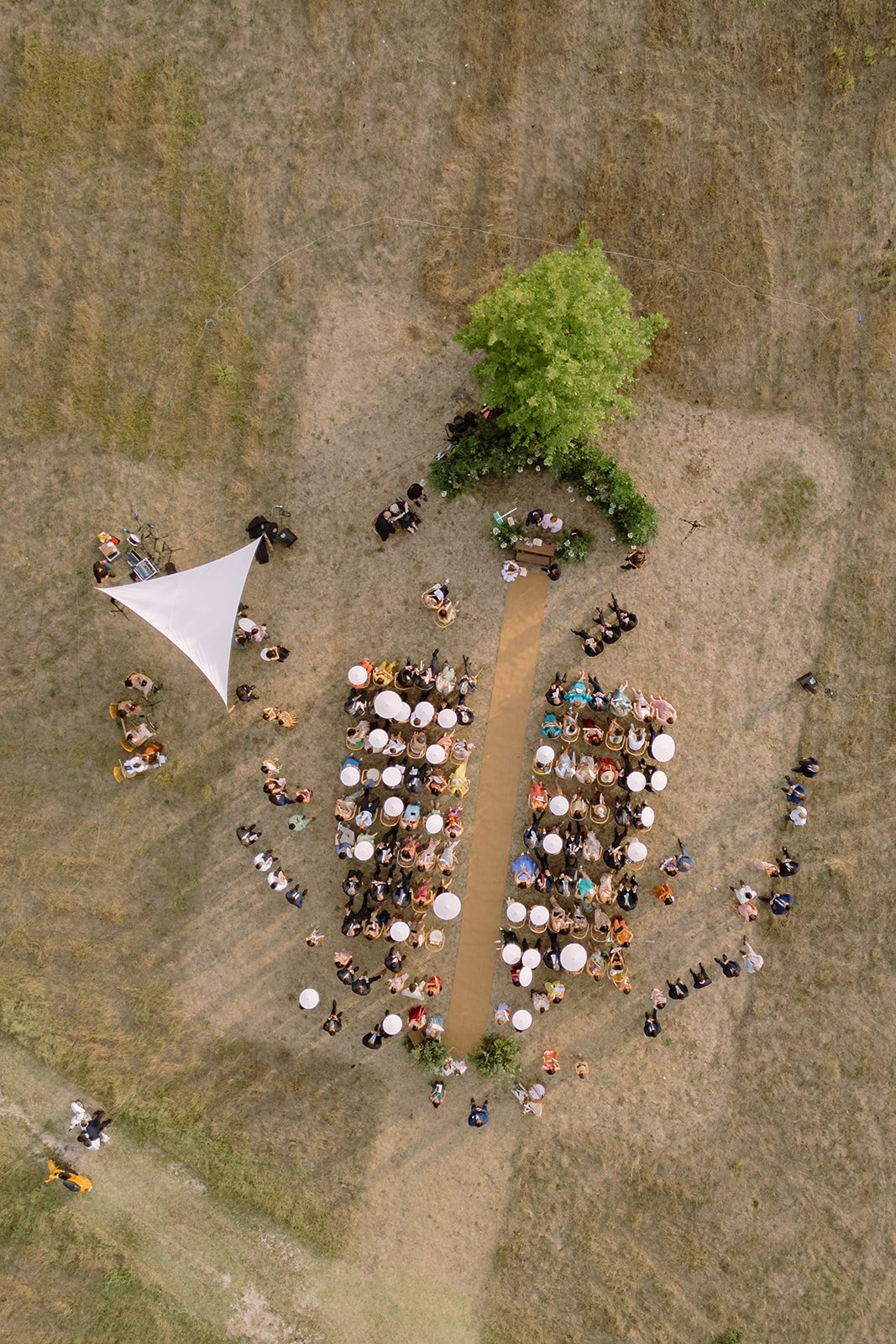 An aerial view of an outdoor event with a large group of people gathered around tables on a grassy area, a pathway leading to a tree with chairs and a table underneath, and some tents with equipment nearby.