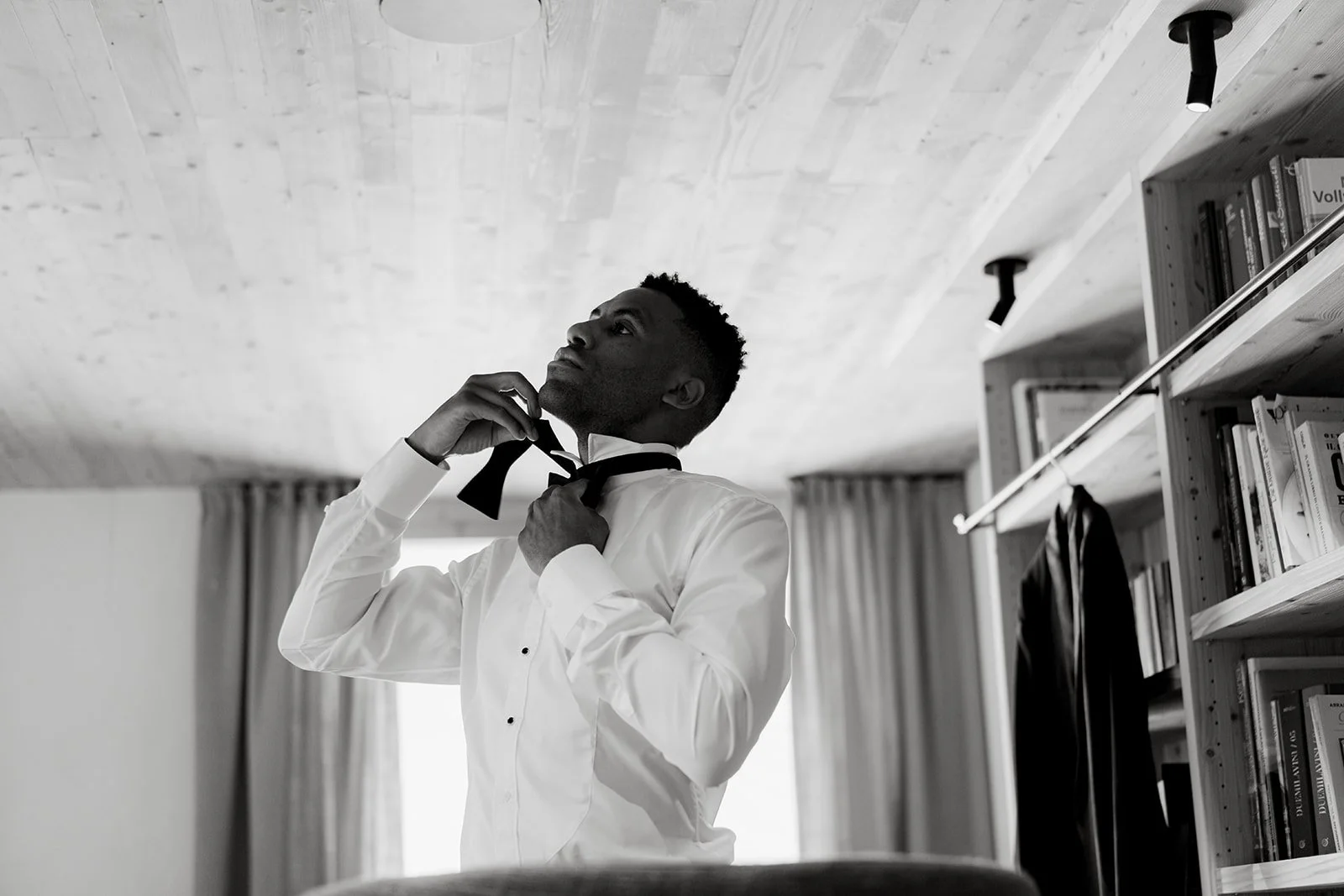 A man in a white shirt is adjusting a black bowtie in a room with wooden ceiling and shelves with books.