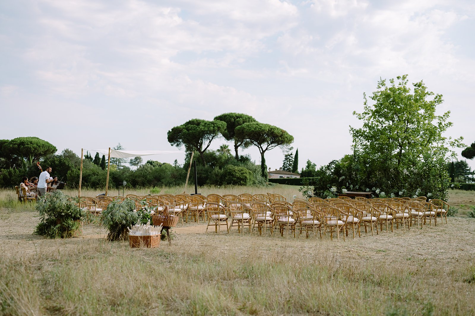 Outdoor wedding ceremony setup with chairs arranged in rows, large trees in the background, and a few people preparing on the side.