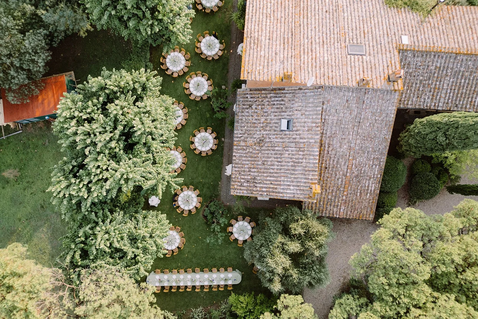 Overhead view of a garden with multiple round tables covered with white tablecloths, surrounded by chairs, set up for an outdoor event or celebration, with green trees and a building with tiled roofs nearby.