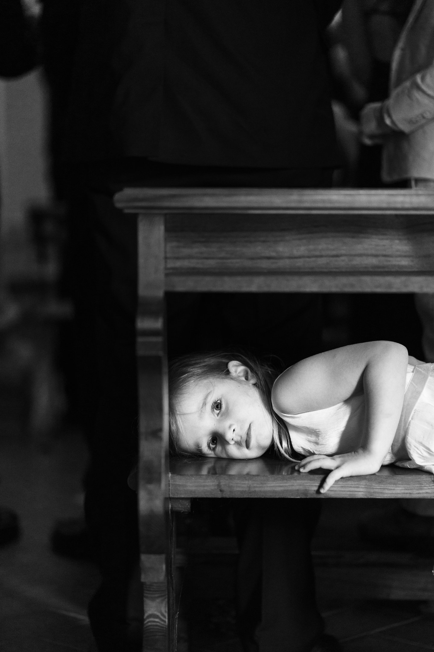 A young girl lying on her side under a wooden table, looking directly at the camera with a contemplative expression in a black and white photo.