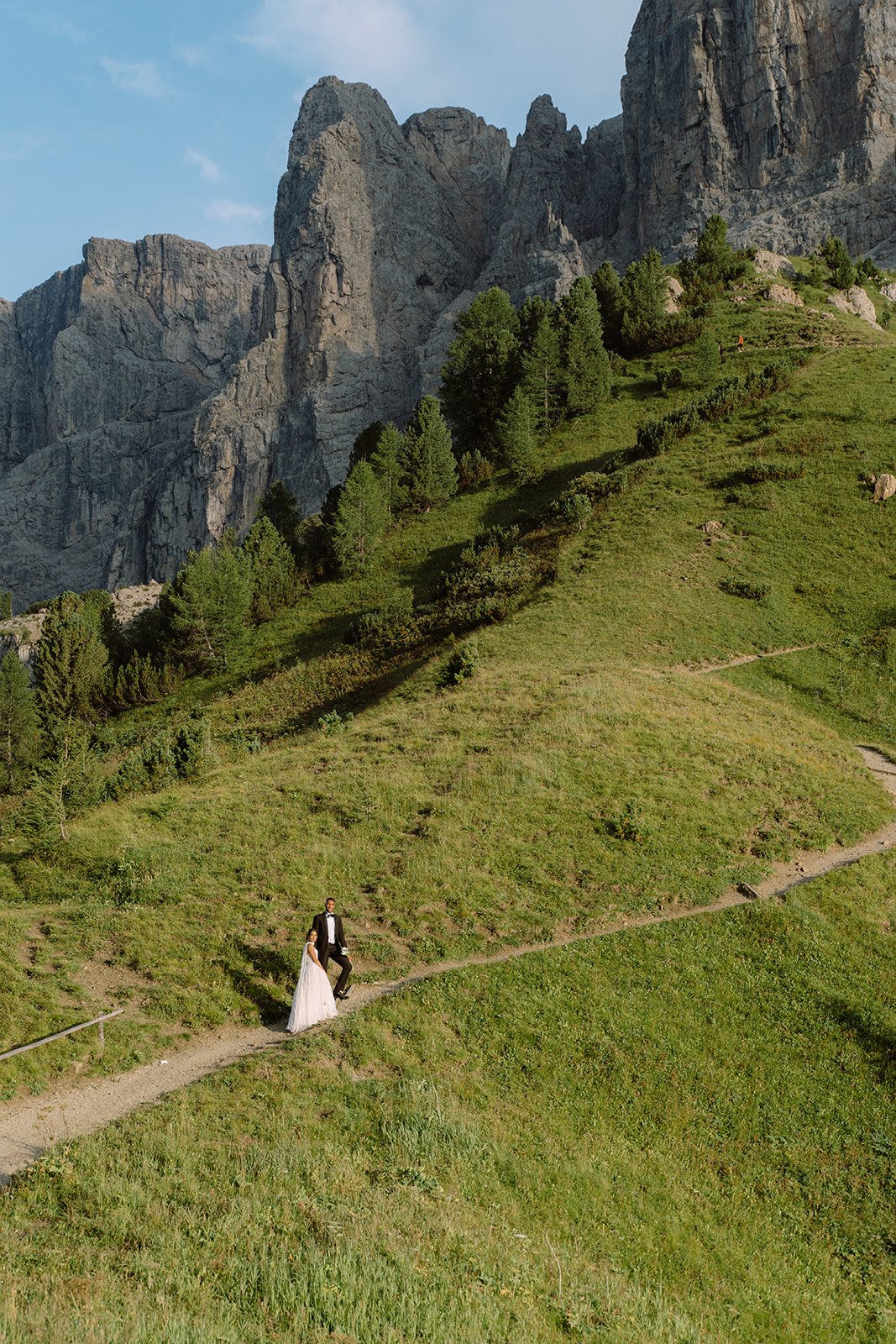 A bride and groom walking on a narrow dirt path in a lush green hillside with tall rocky mountains in the background.
