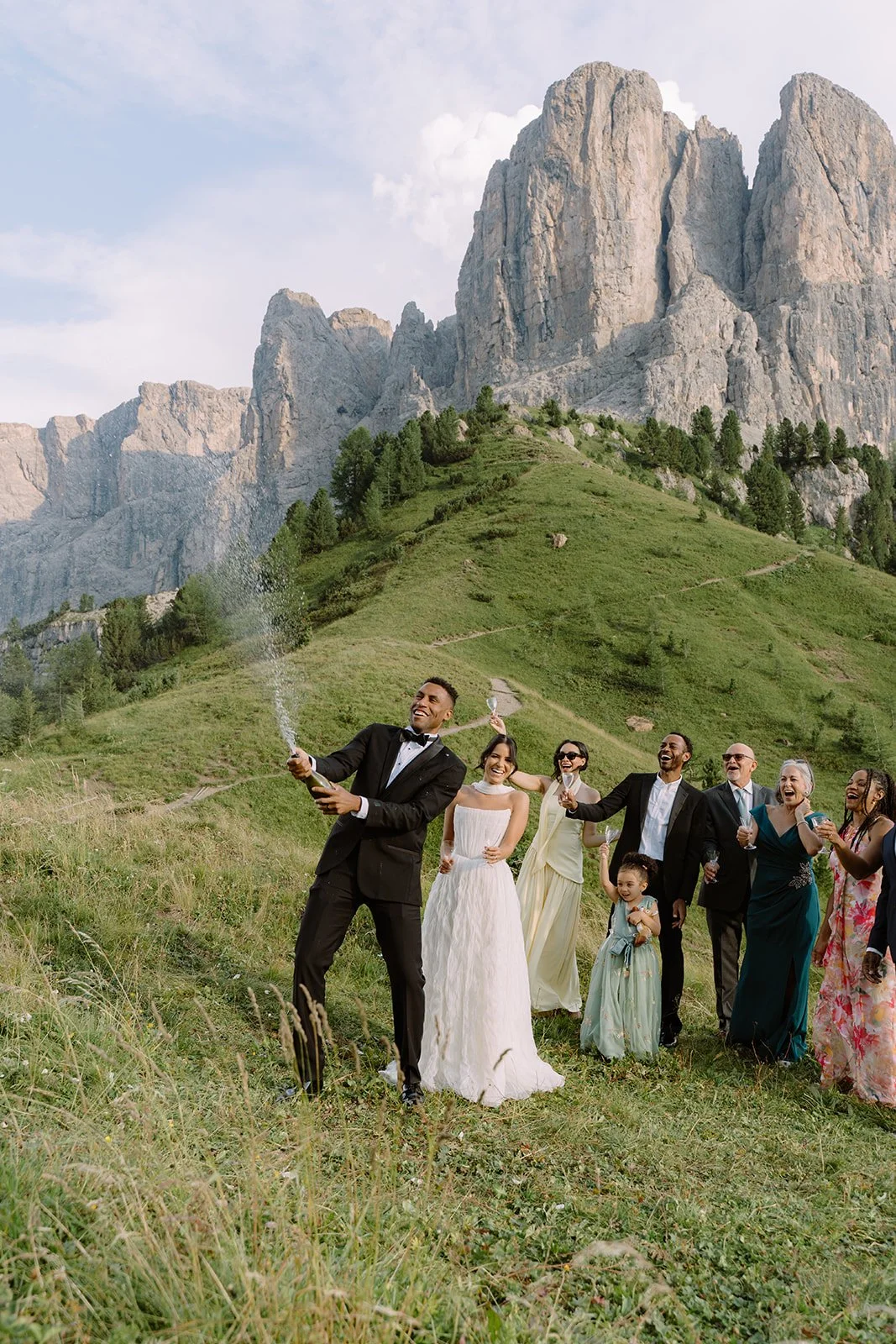 A wedding celebration outdoors with a diverse group of people in formal attire, celebrating together in a mountain meadow with rocky peaks and green hills in the background.