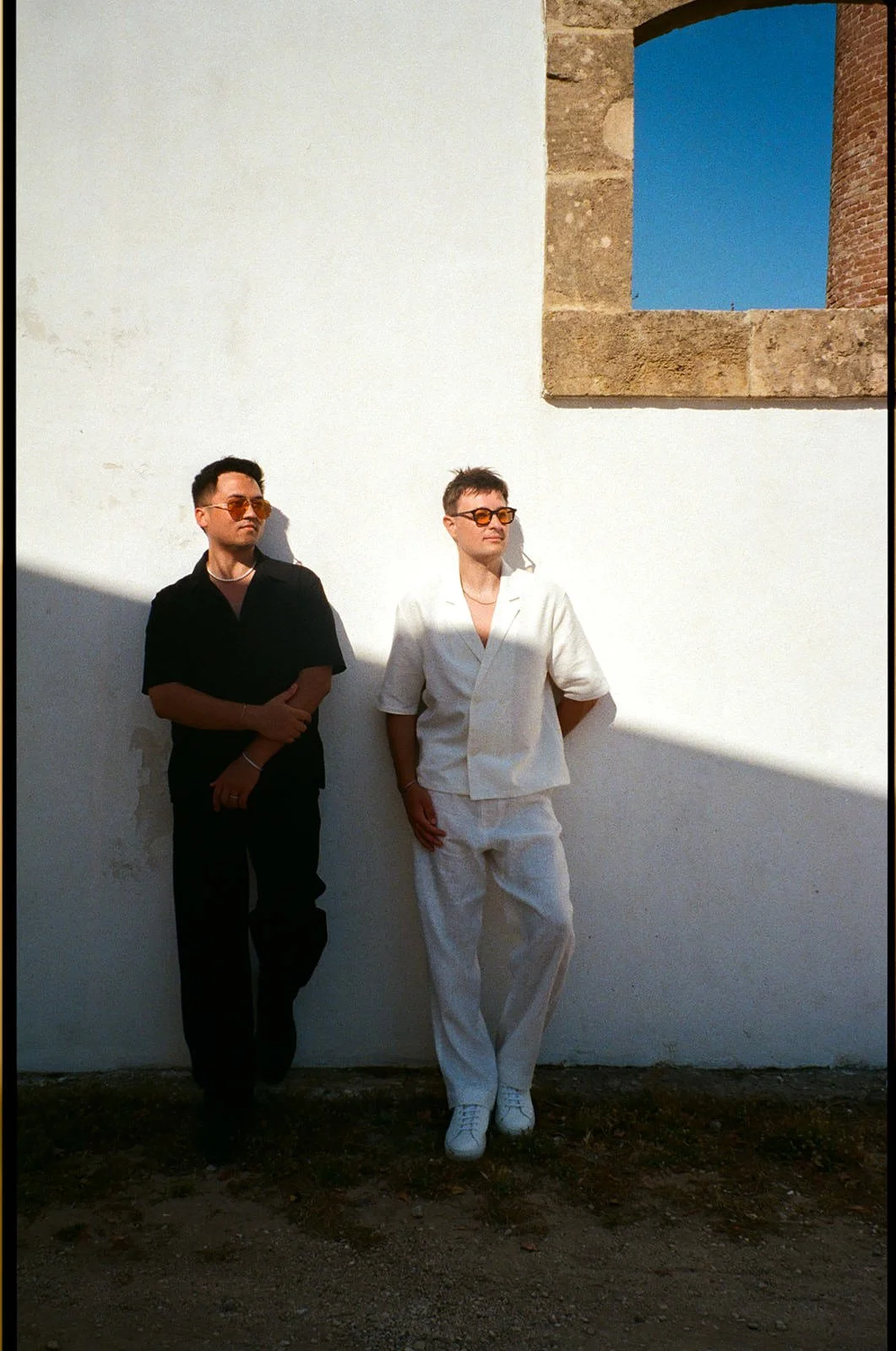 Two people, one in black and one in white, standing against a white wall with a small window, wearing sunglasses, with shadows cast on the wall.