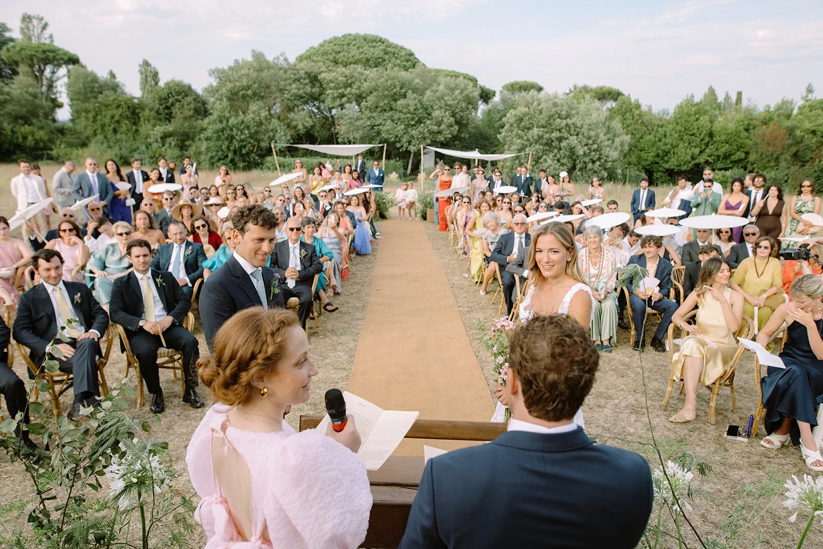 Outdoor wedding ceremony with a large seated crowd, officiant reading at a wooden podium, bride and groom at the altar, and guests seated on both sides of an aisle, with trees in the background.