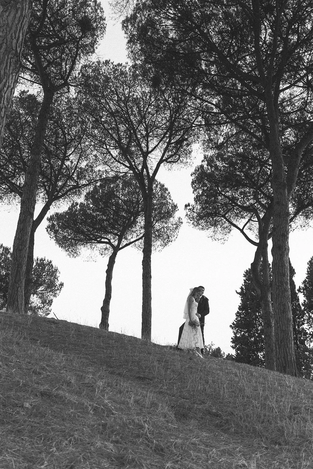 A black-and-white photo of a couple in wedding attire embracing on a grassy hillside surrounded by tall trees.