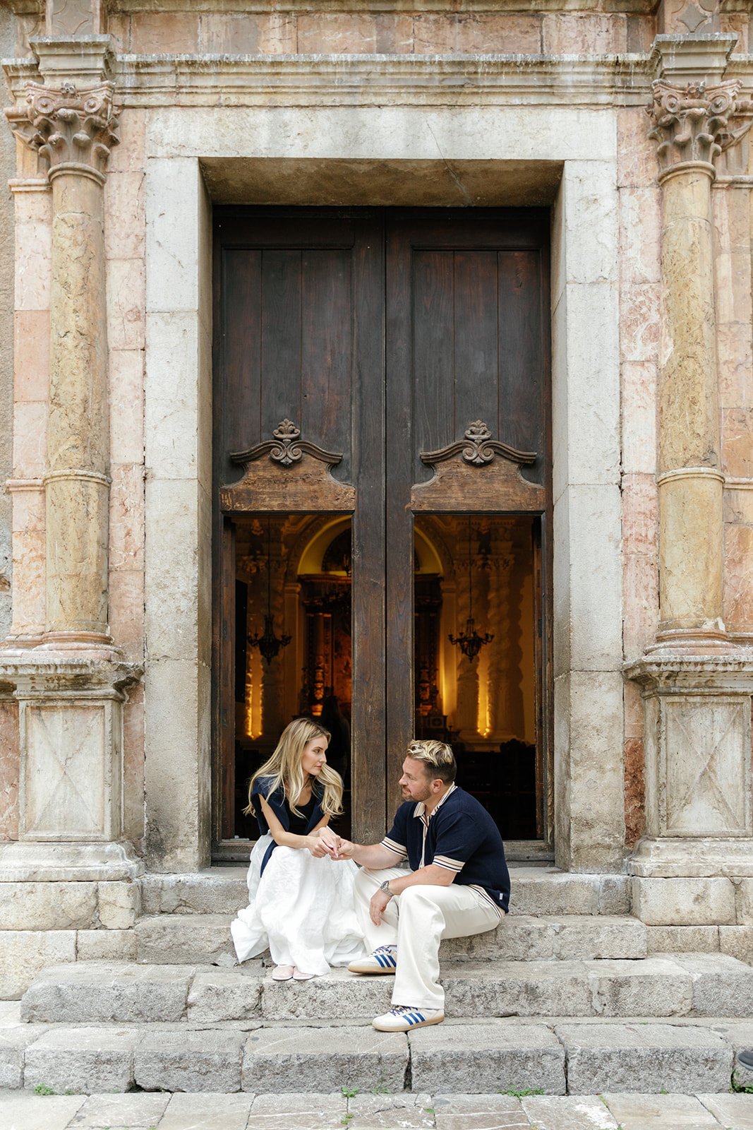 A young woman and a young man sitting on stone steps outside a historic building with large wooden door, holding hands and engaging in conversation.