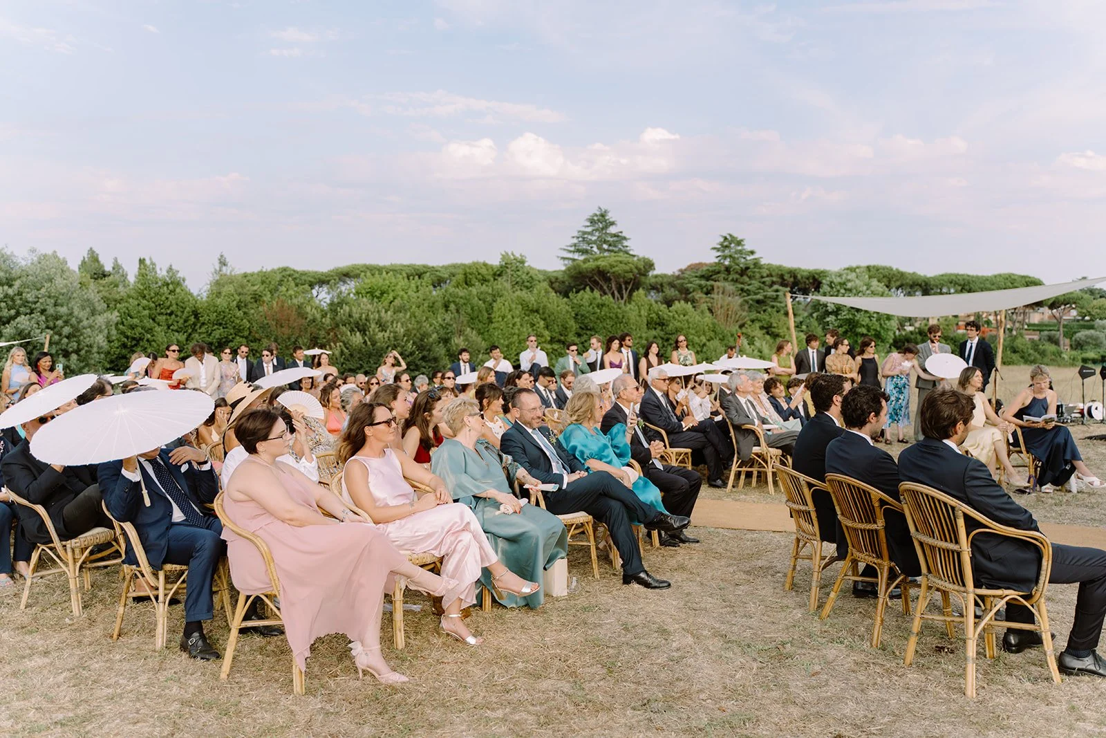 A large outdoor wedding ceremony with guests sitting on wooden chairs, some holding white umbrellas, on a grassy area under a sunny sky with scattered clouds, surrounded by green trees.