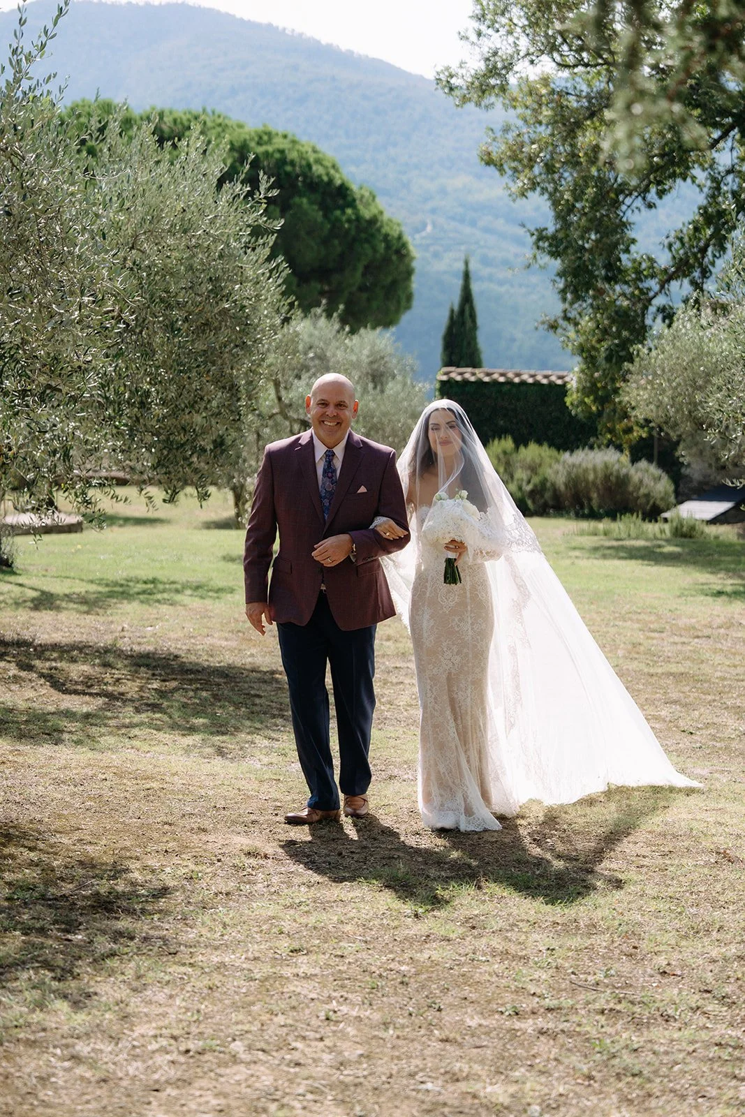 A bride walking down an outdoor aisle with her father in a scenic garden setting with trees and mountains in the background.