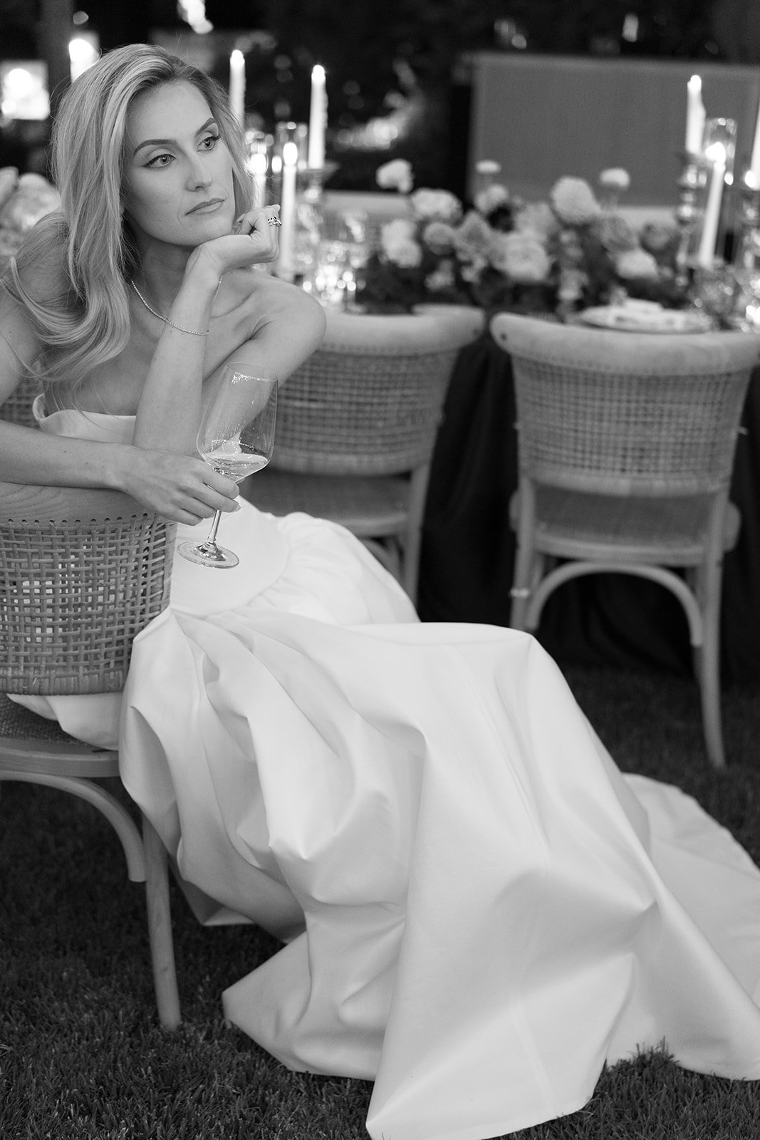 A woman in a strapless dress sits on a chair at a dinner event, holding a wine glass, with a thoughtful expression, surrounded by decorated tables with flowers and candles.