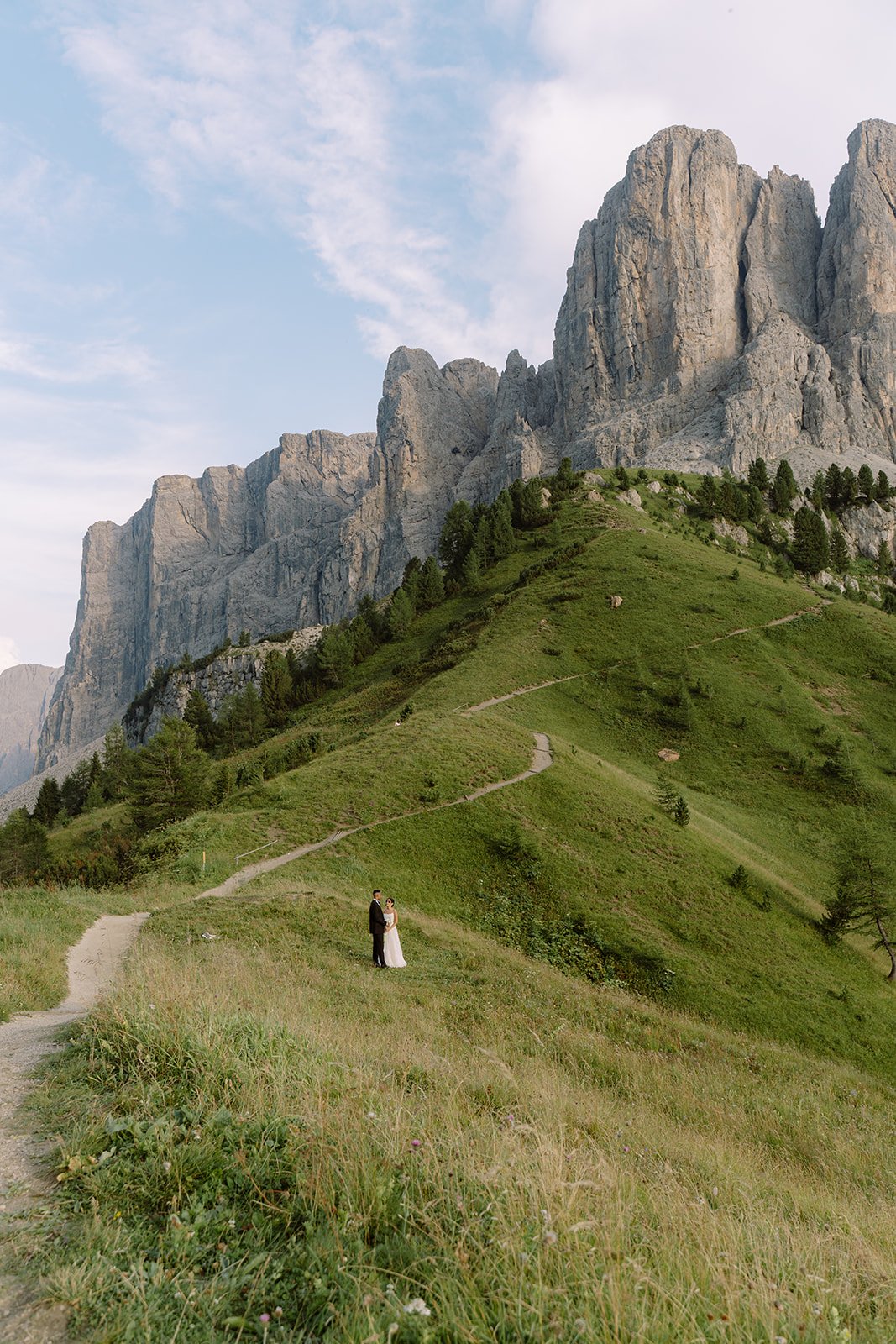 A bride and groom standing on a grassy hillside with mountain peaks in the background.