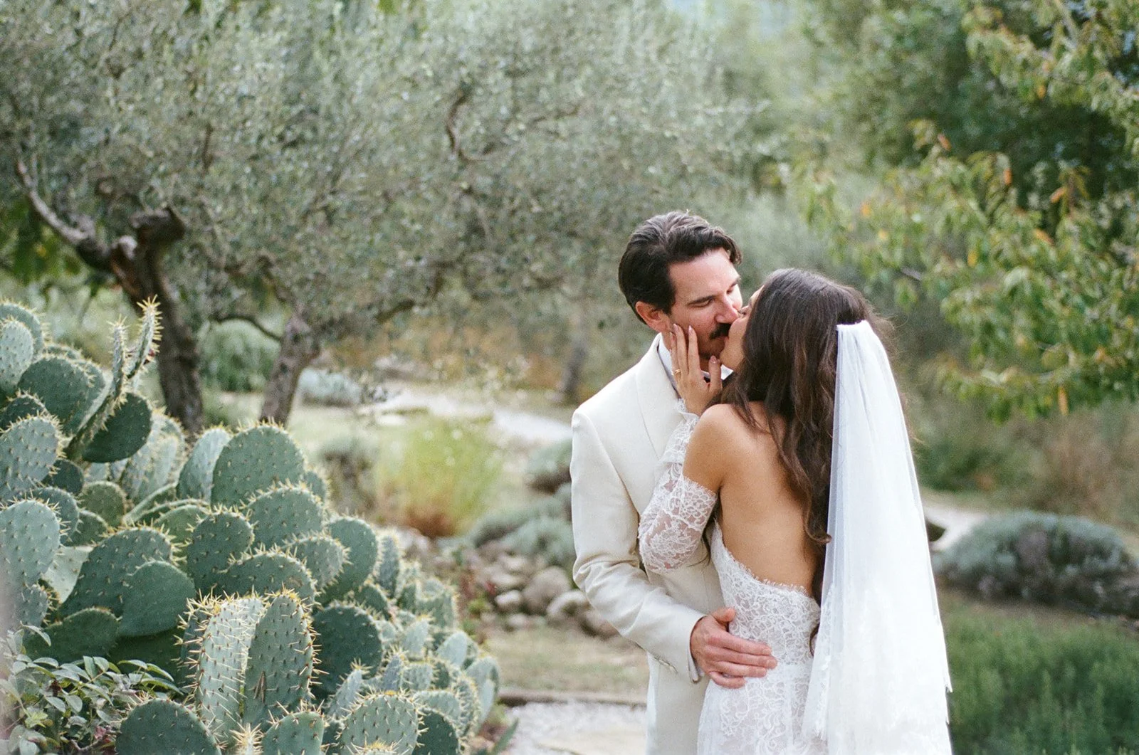 A bride and groom share a kiss outdoors in a garden with greenery and cacti.