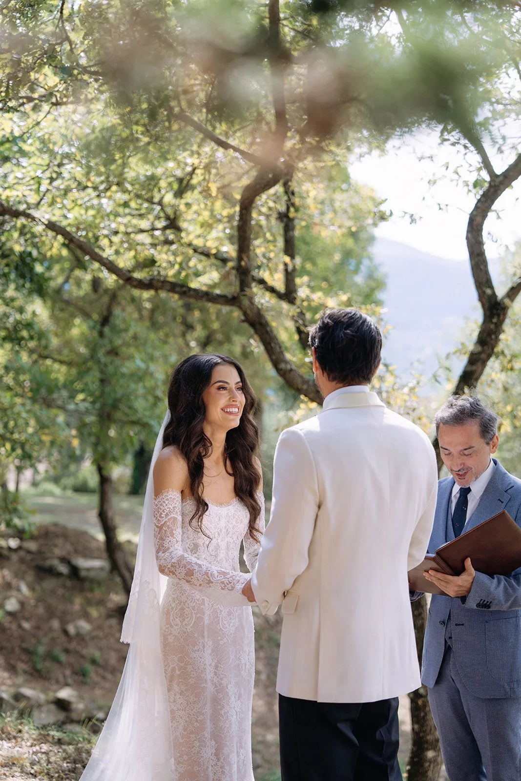 A bride and groom holding hands during their outdoor wedding ceremony, with a officiant reading from a book, under trees in a natural setting.
