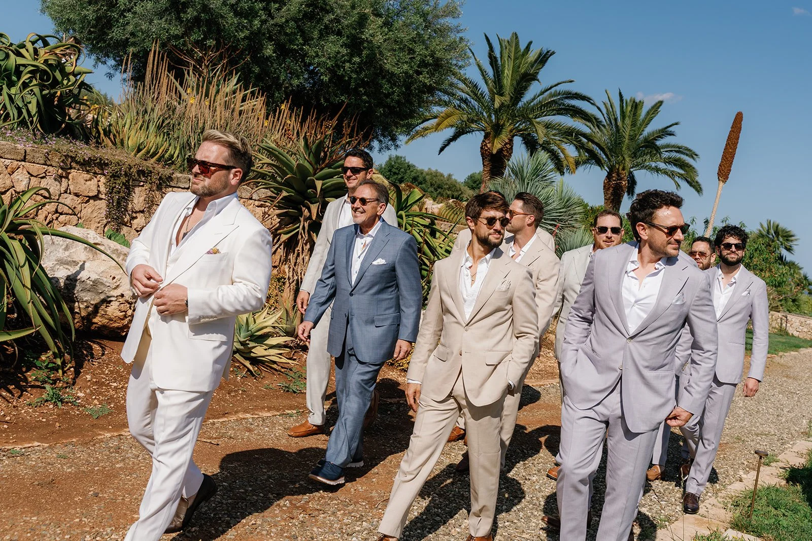 A group of men in light-colored tuxedos and suits walking outdoors on a bright, sunny day, with palm trees and desert plants in the background.