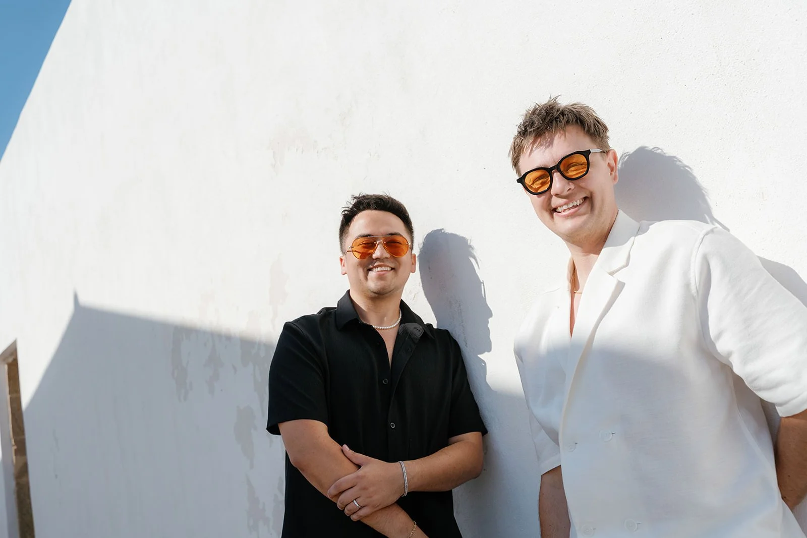 Two men smiling and leaning against a white wall, wearing sunglasses, one in black shirt and the other in white blazer, during daytime.