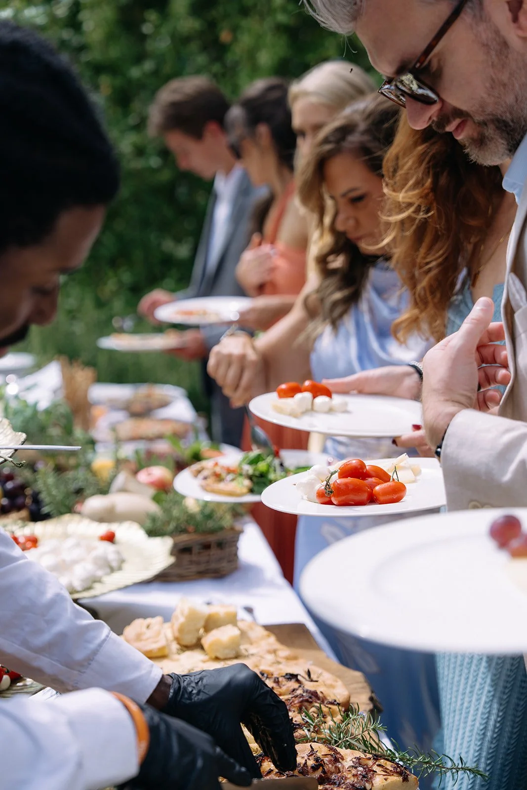 People in line at an outdoor buffet with various dishes, including tomatoes, cheese, bread, and grilled meat, in a lush green setting.