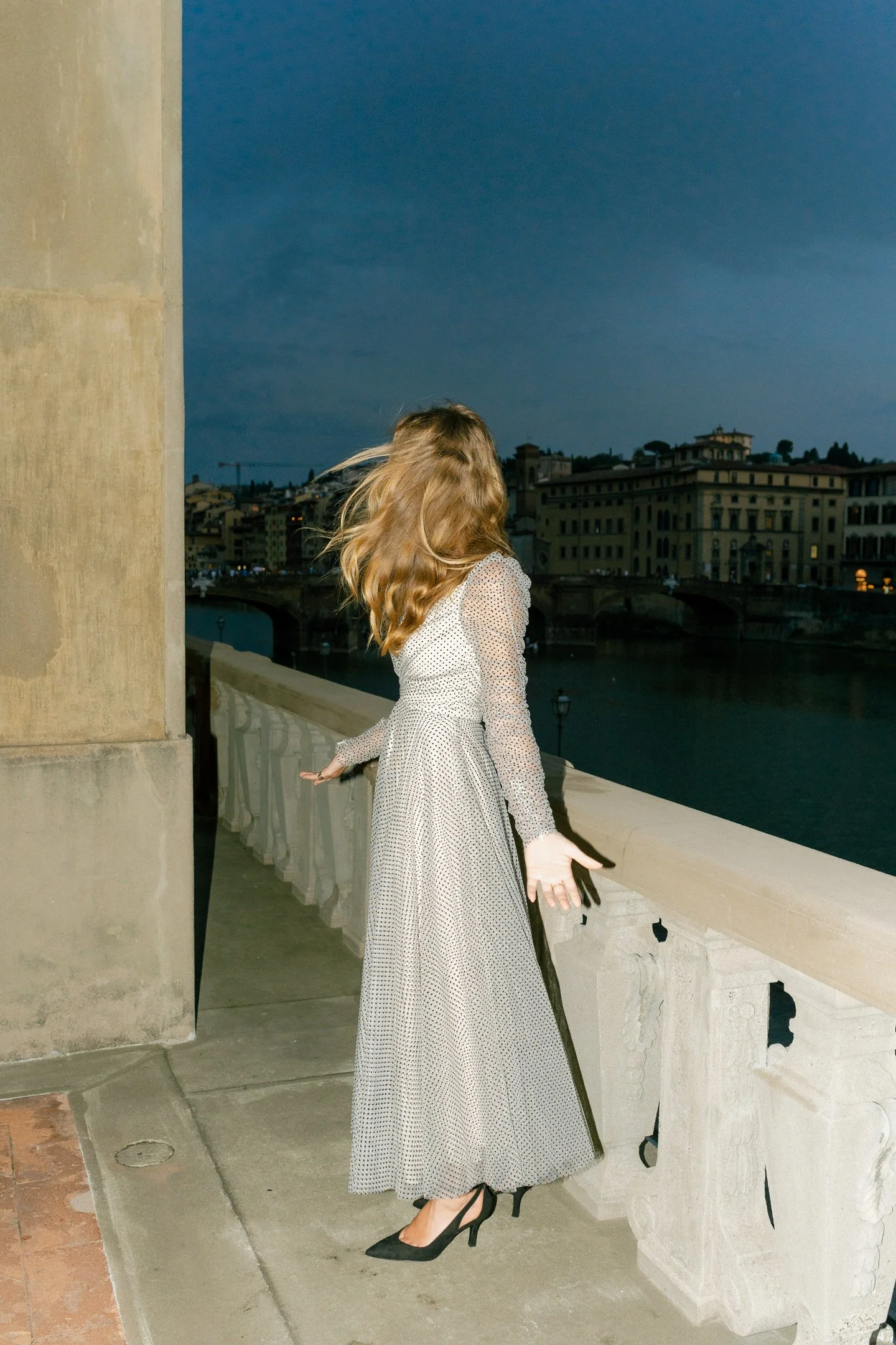 A woman with wavy blonde hair wearing a long, patterned dress and black high heels stands on a bridge over a river at dusk, with a cityscape and buildings in the background.